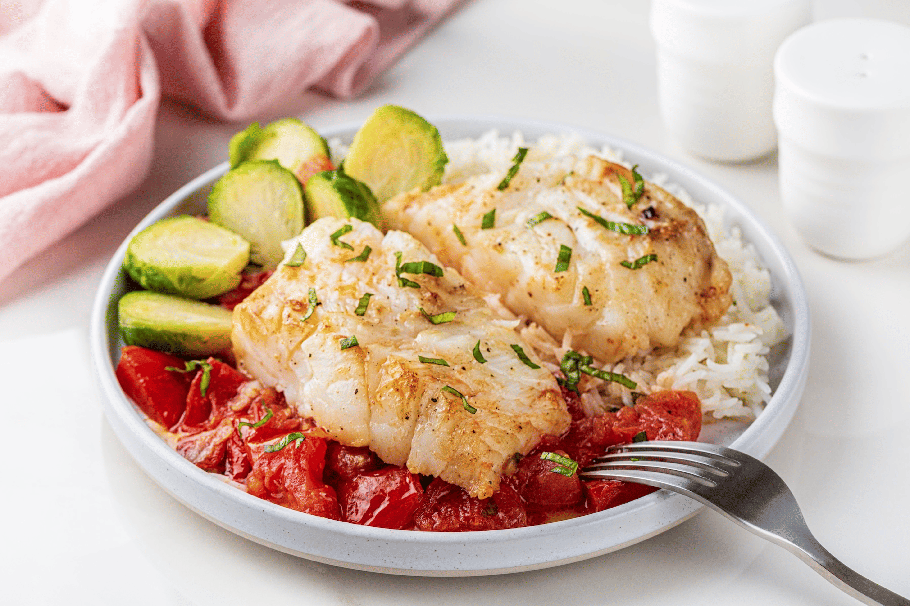 A plate of cod fish served with tomatoes, Brussel sprouts, and rice.