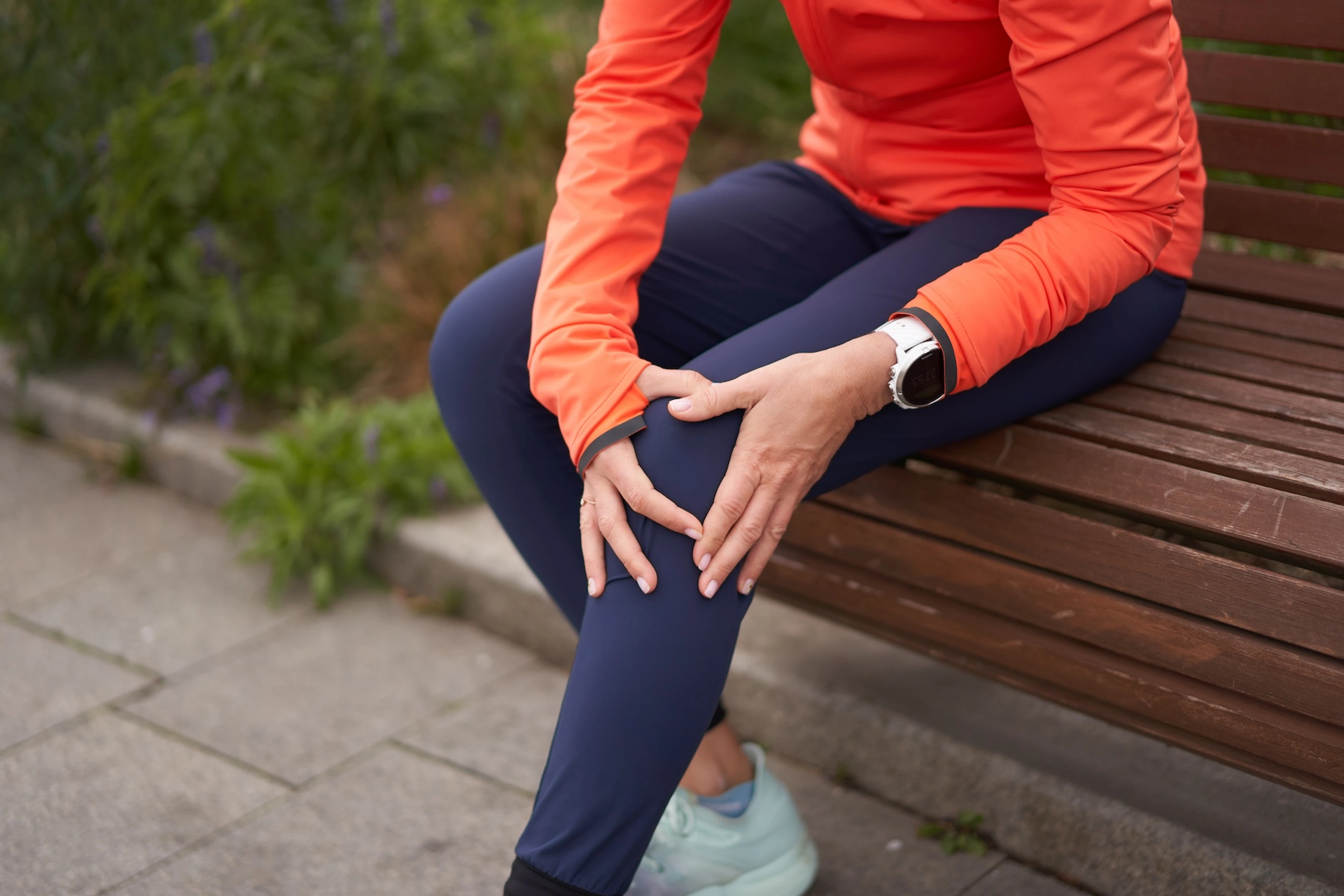 A woman experiencing runner's knee. It's a close-up photo of her sitting on a park bench after a run and holding onto the front of her left knee.
