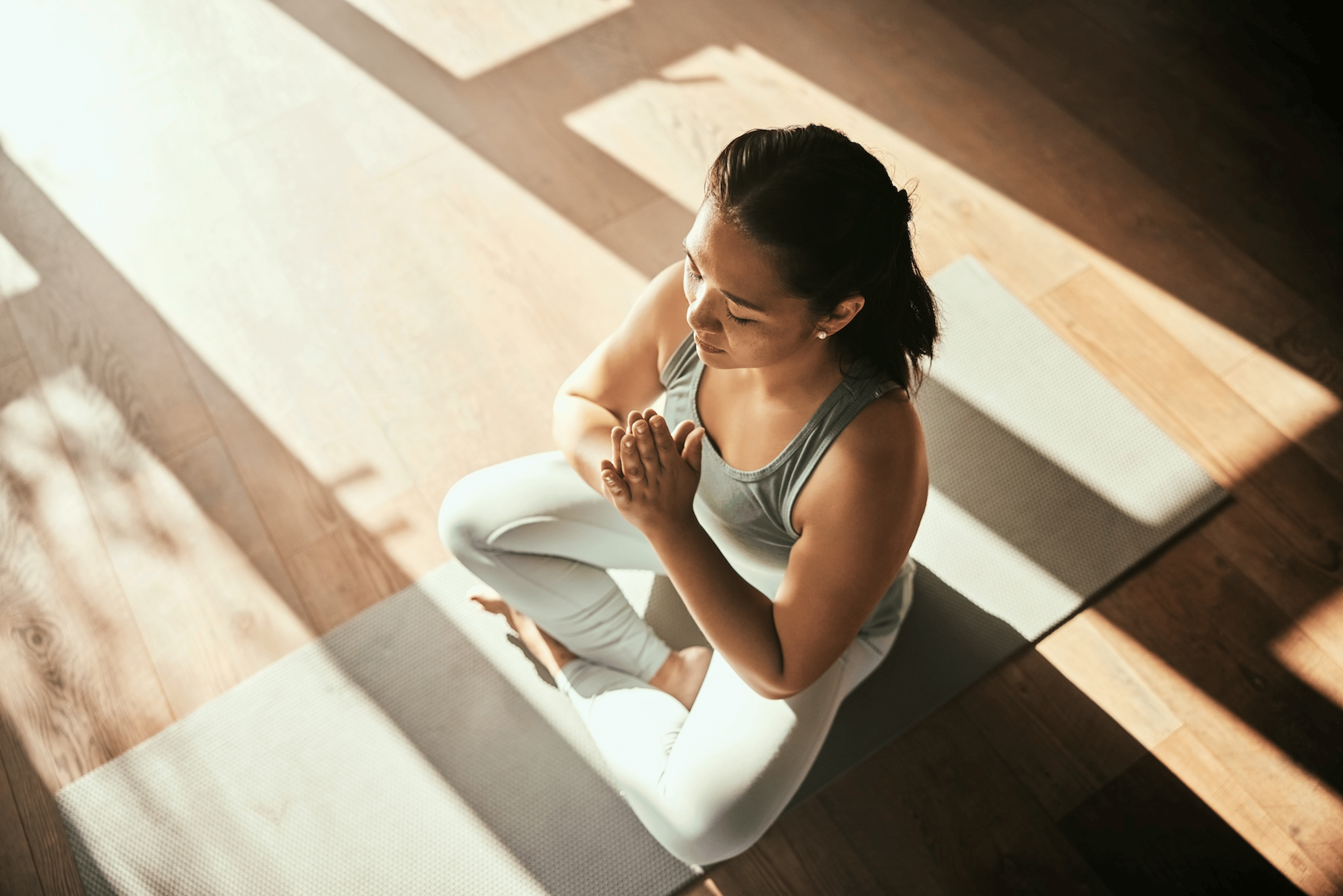 A woman practicing self-care by meditating at home on a yoga mat in the sunlight.