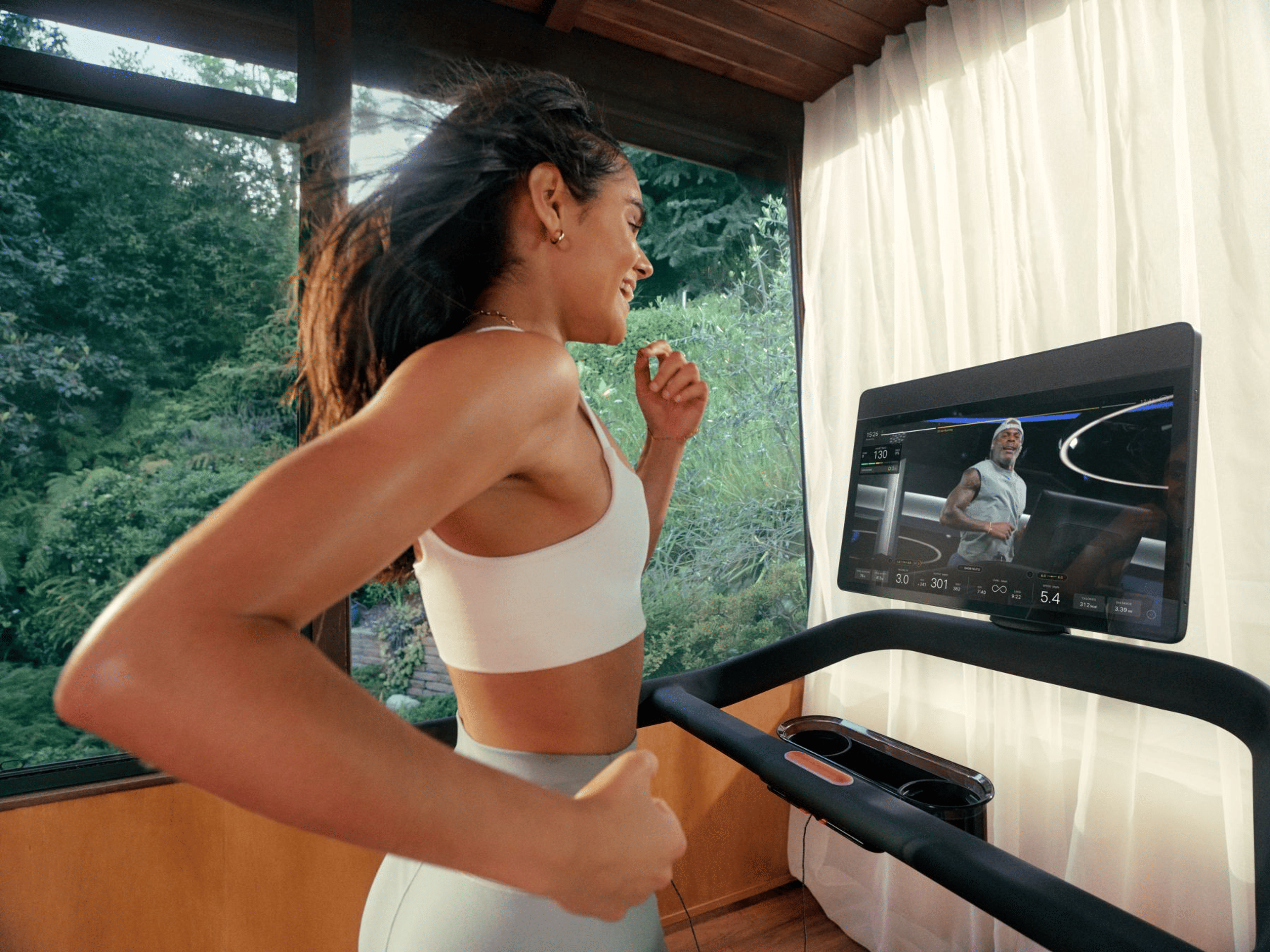 A person is running on a treadmill with a screen that displays a virtual coach. The person is smiling brightly and looks happy and their arms are swinging and hair blowing back as they run.