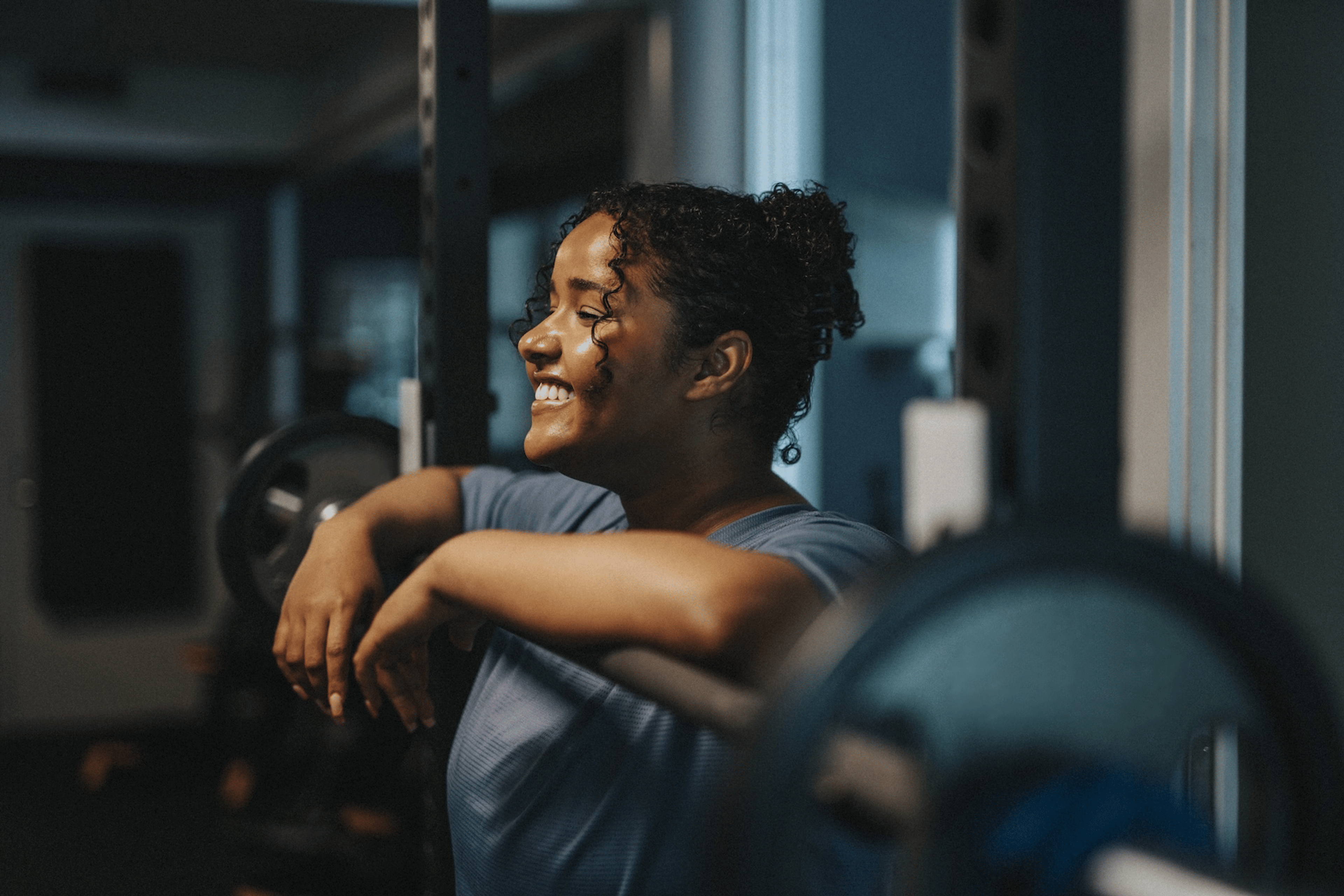 A woman smiling and resting her arms on a barbell. She is enjoying her exercise routine and views working out as a hobby.