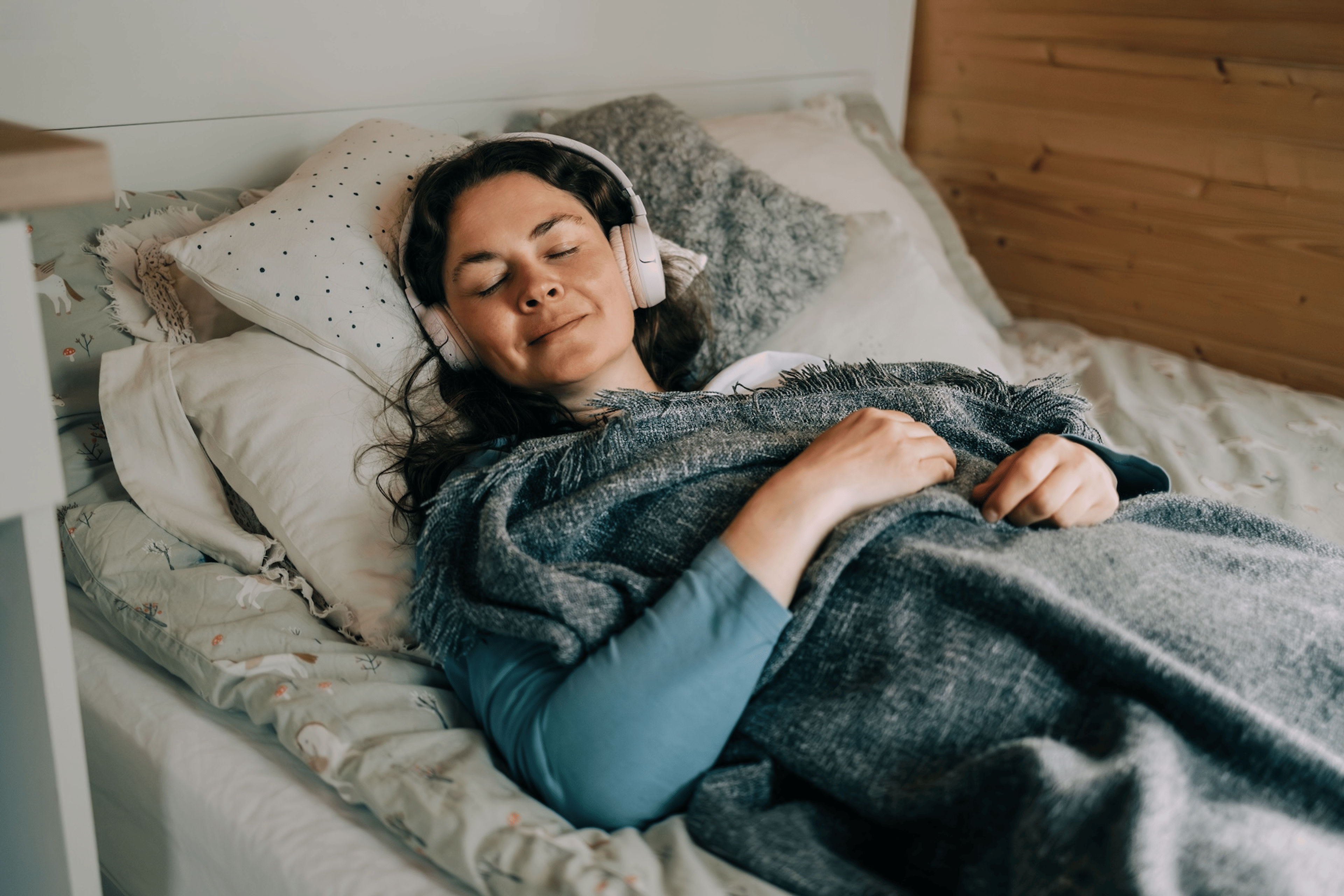 A relaxed woman meditating in bed. She has headphones on and is under a blanket.