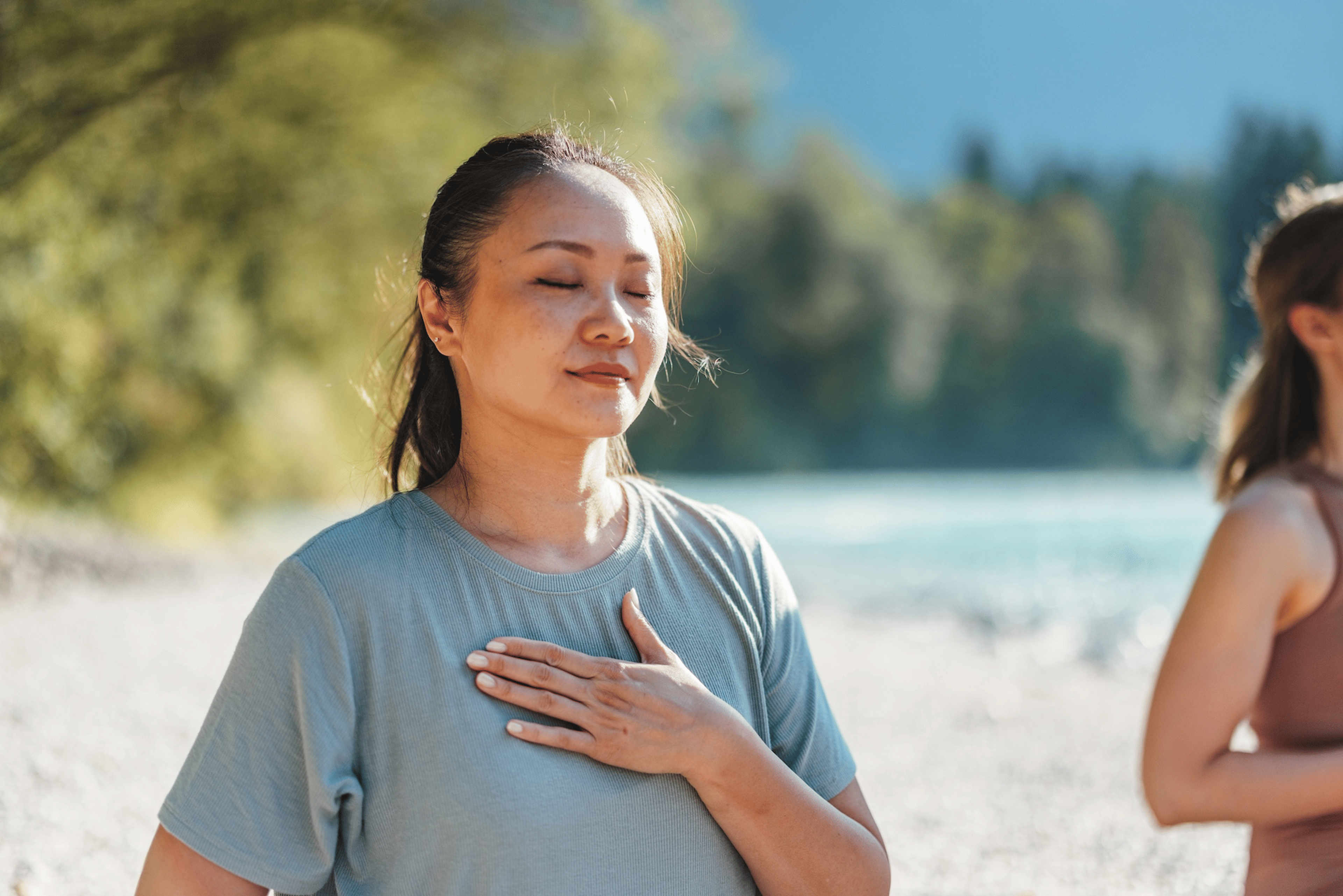 A woman practicing self-compassion meditation outside, softly smiling and holding her hand to her heart.