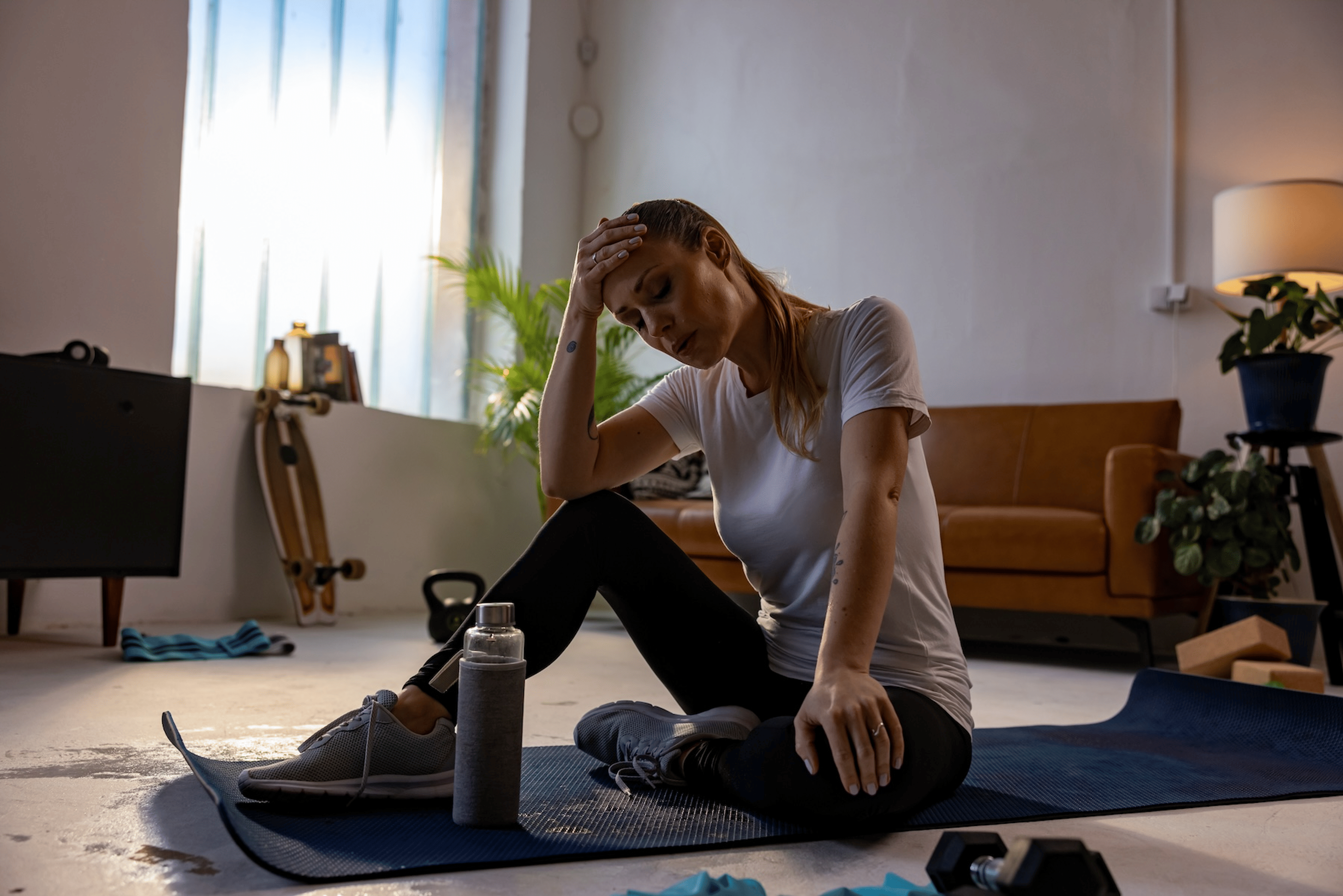 A woman sitting down on a yoga mat at home with her head resting in her hands and her eyes clothes as she takes deep breaths. She felt dizzy when stretching.