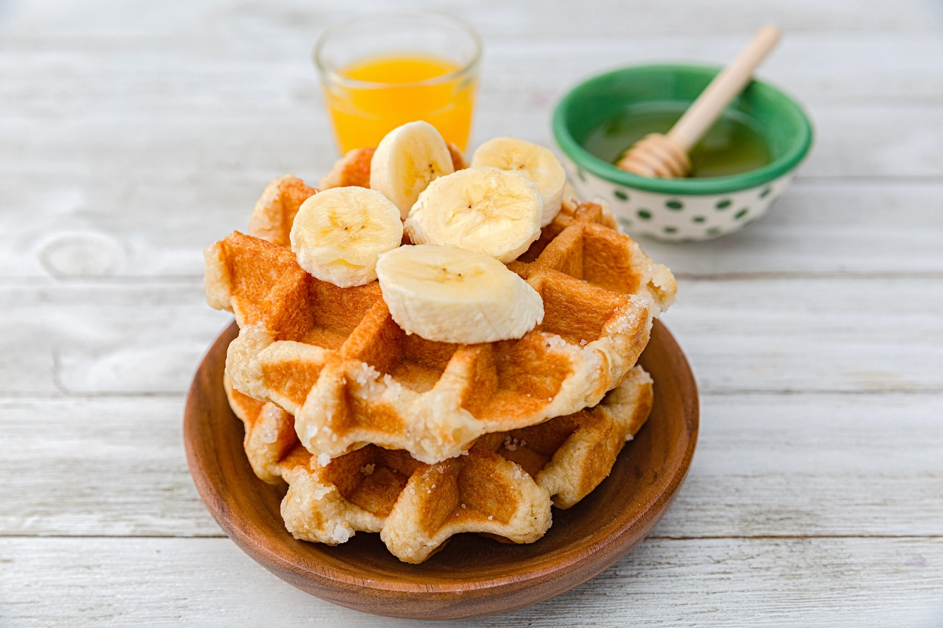 A plate of Belgian waffles with banana slices. There's a bowl of syrup or honey and a glass of juice next to the plate.