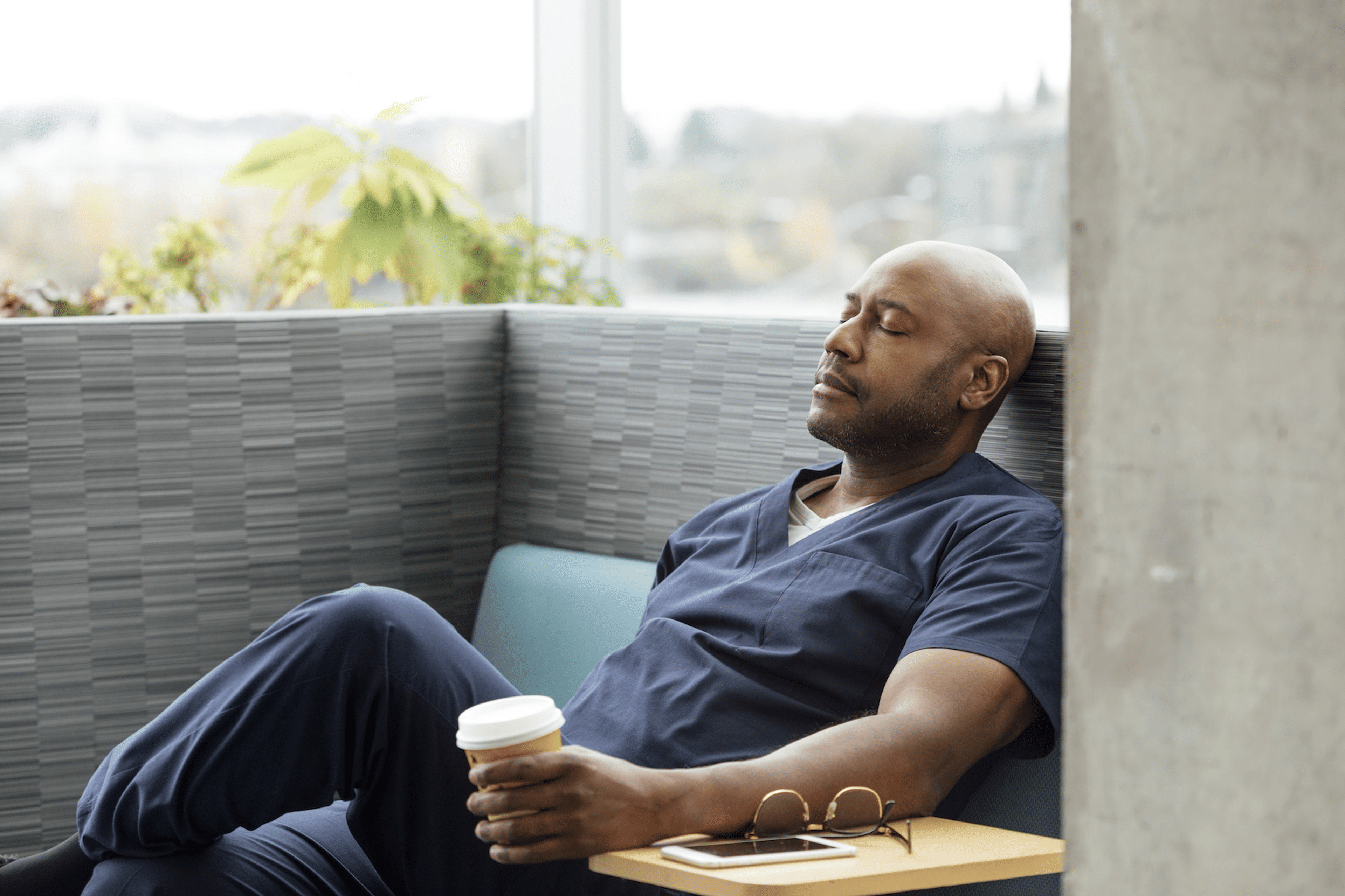 A male healthcare worker sitting asleep and holding a cup of coffee in the middle of the day. He's dealing with circadian misalignment.