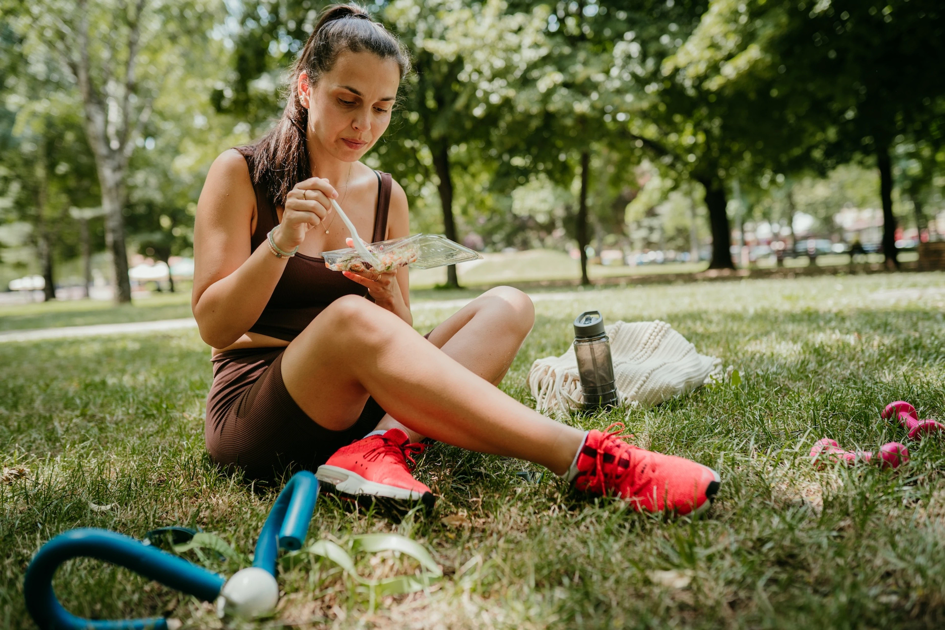A woman eating a meal from a container while sitting in the grass outside. She's wearing workout clothes.