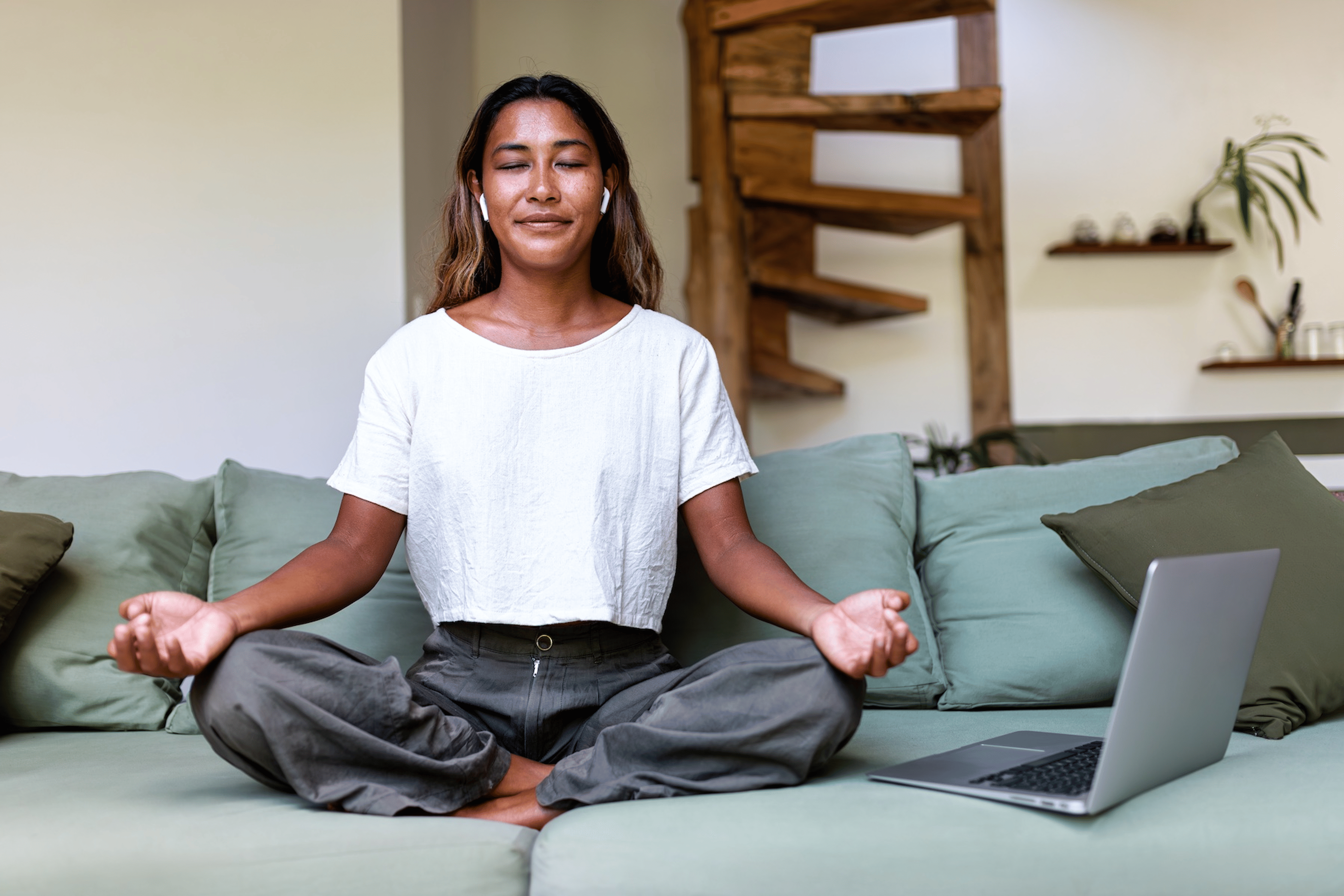 A woman meditating on her couch while listening to a guided meditation on her laptop.