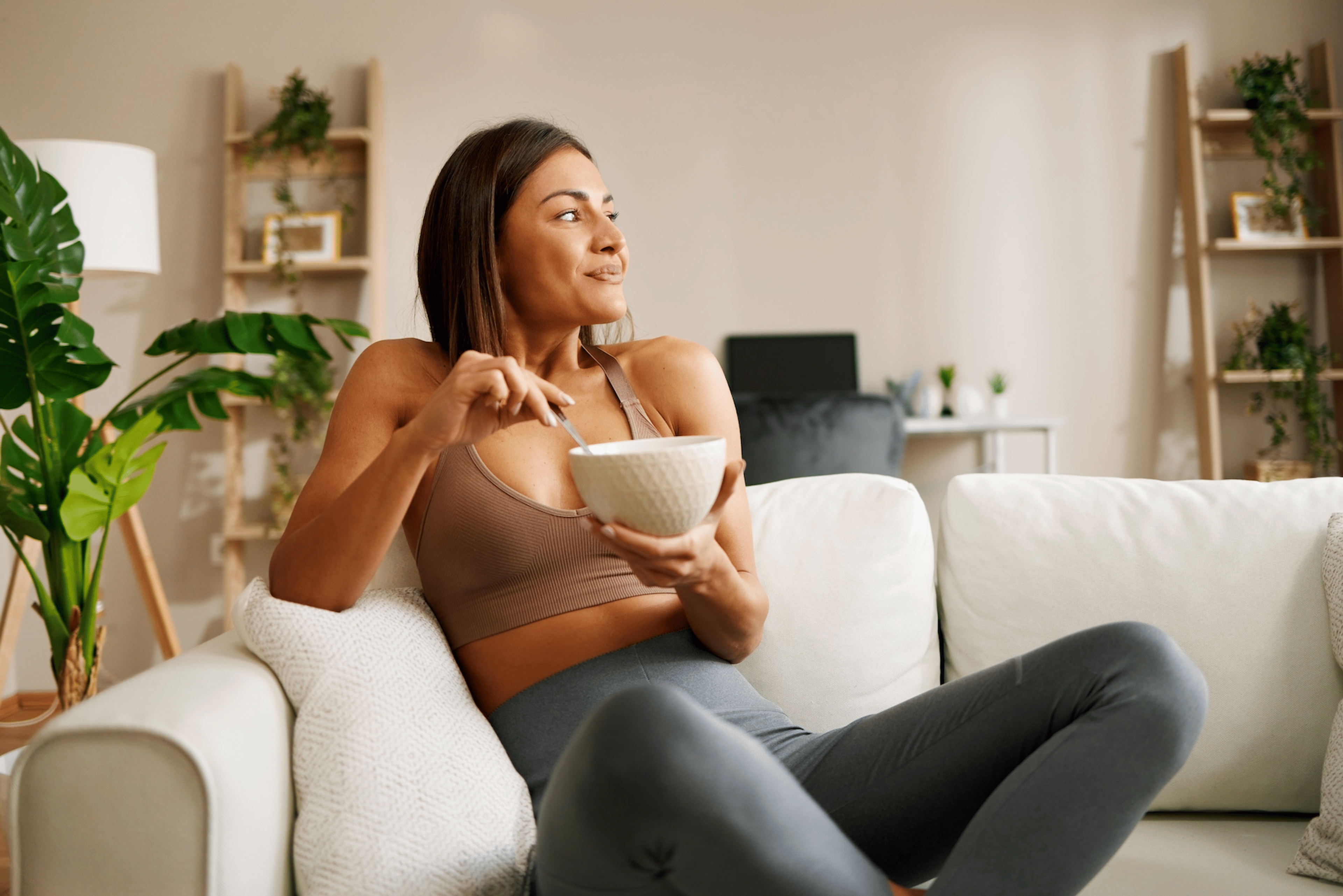 A young woman sitting on the couch at home eating breakfast while wearing workout clothes.