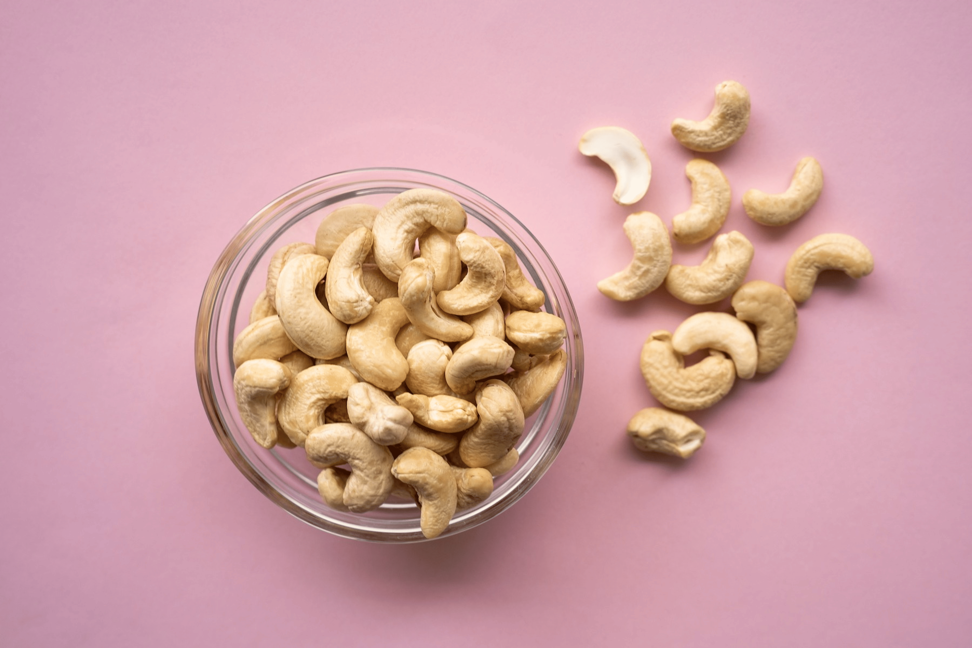 A glass bowl of cashews with some cashews spilled around it on a pink background.