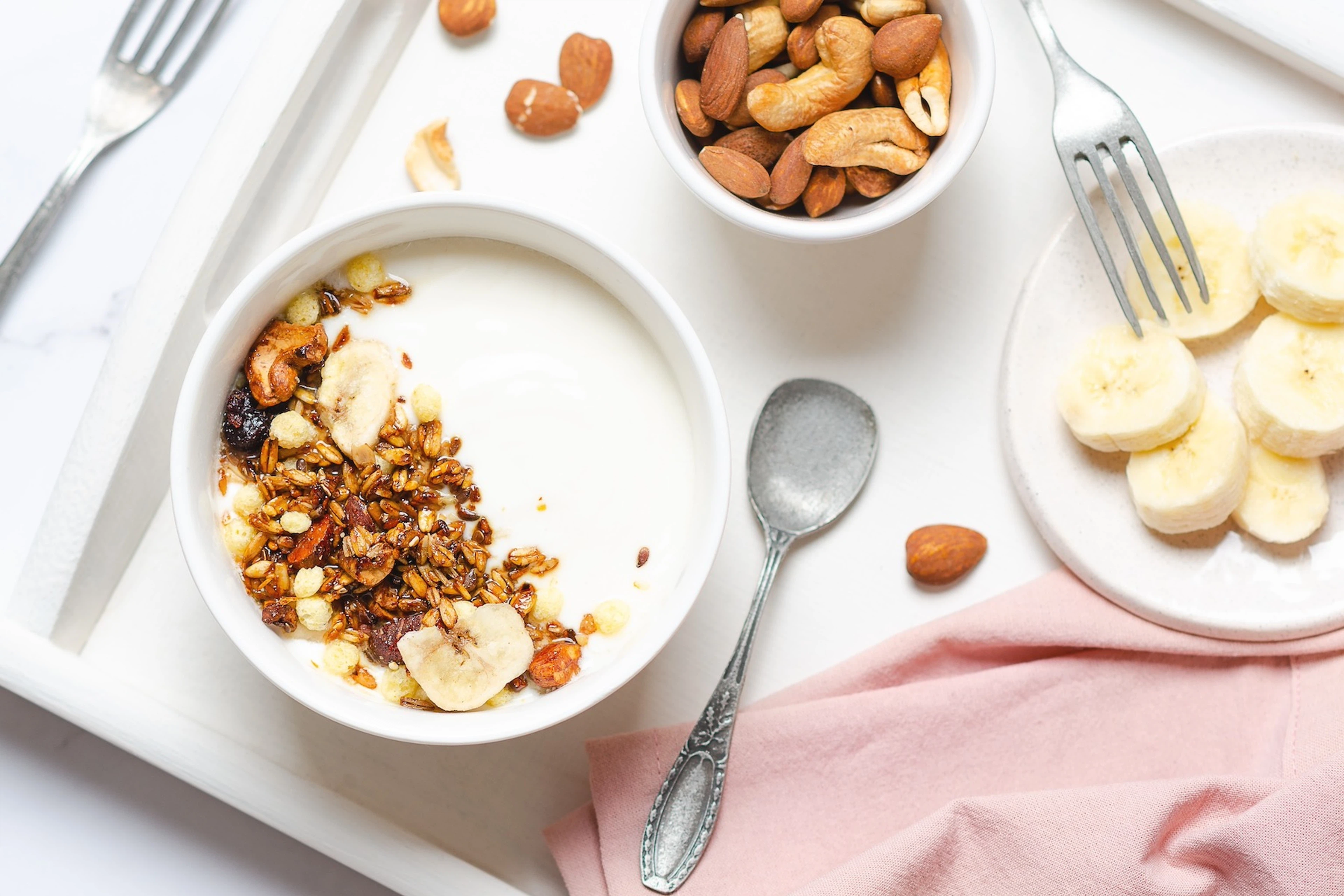 An array of high-protein snacks on a white table, such as Greek yogurt, bananas, and nuts.