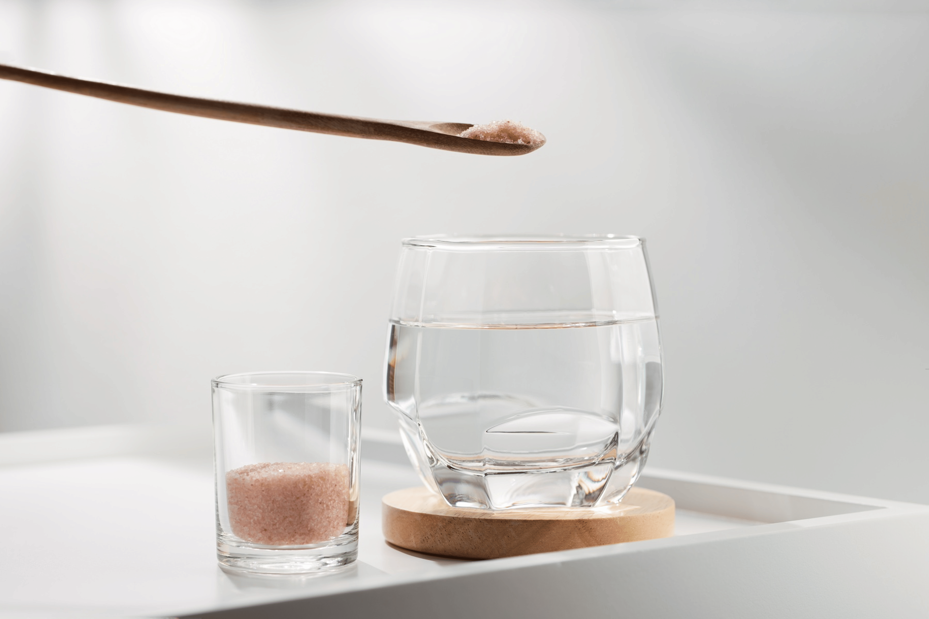 A tiny wooden spoon of pink Himalayan salt hovering over a glass of drinking water. There's a small jar of salt next to the glass as well.