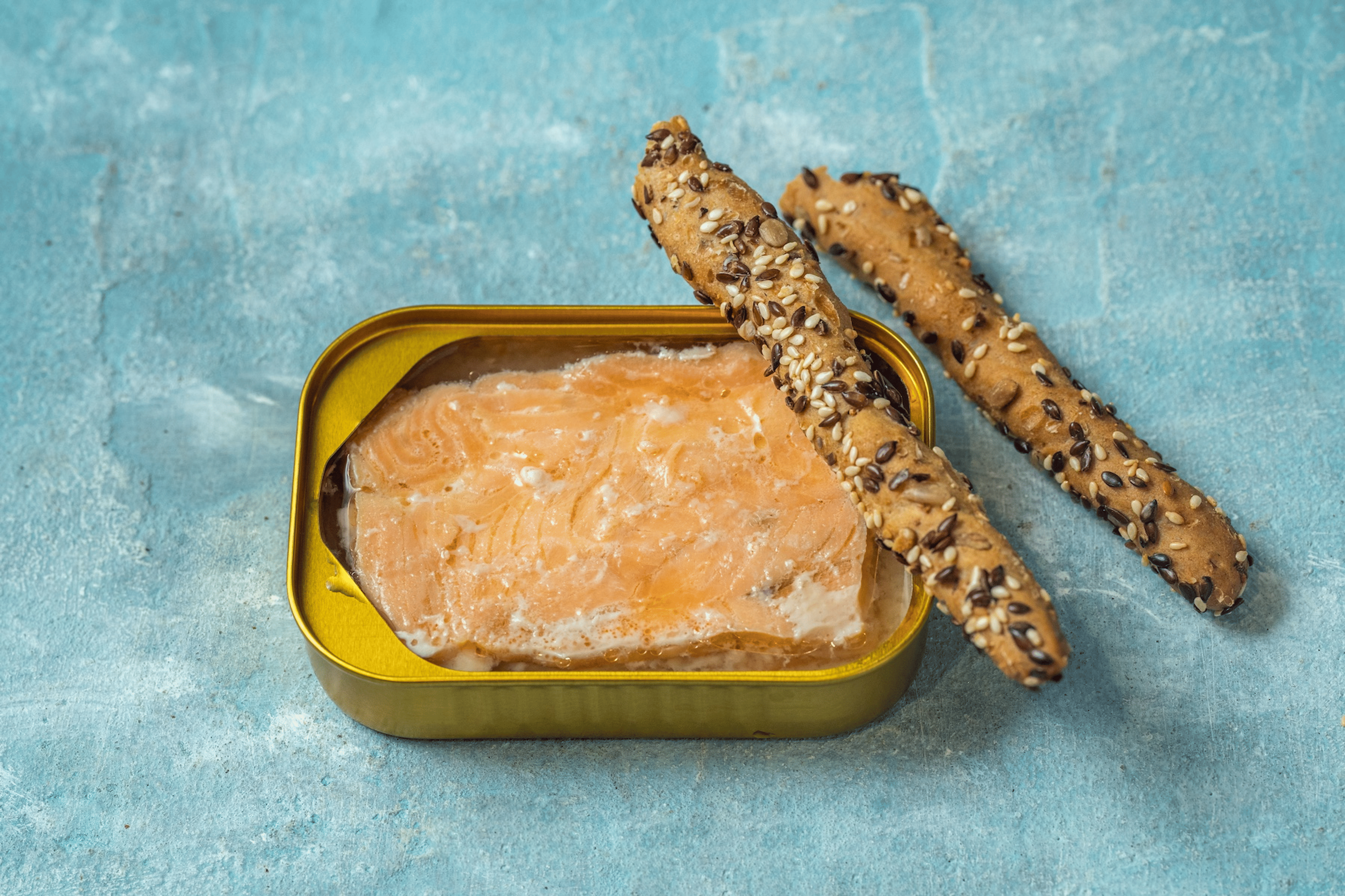 Salmon fillet in an opened tin with two whole-grain breadsticks resting near it.