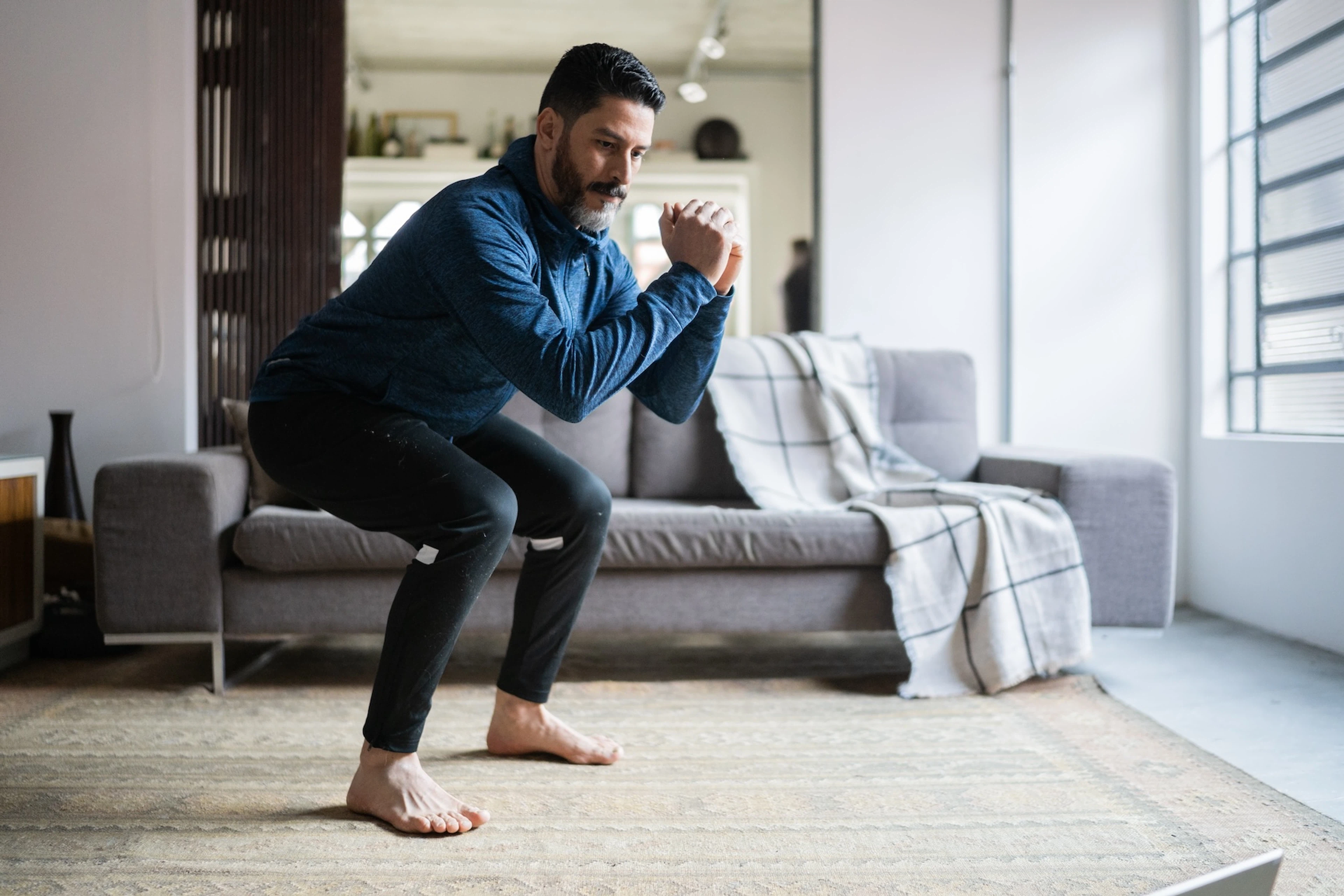A middle-aged man performing a squat exercise while following a guided workout at home.