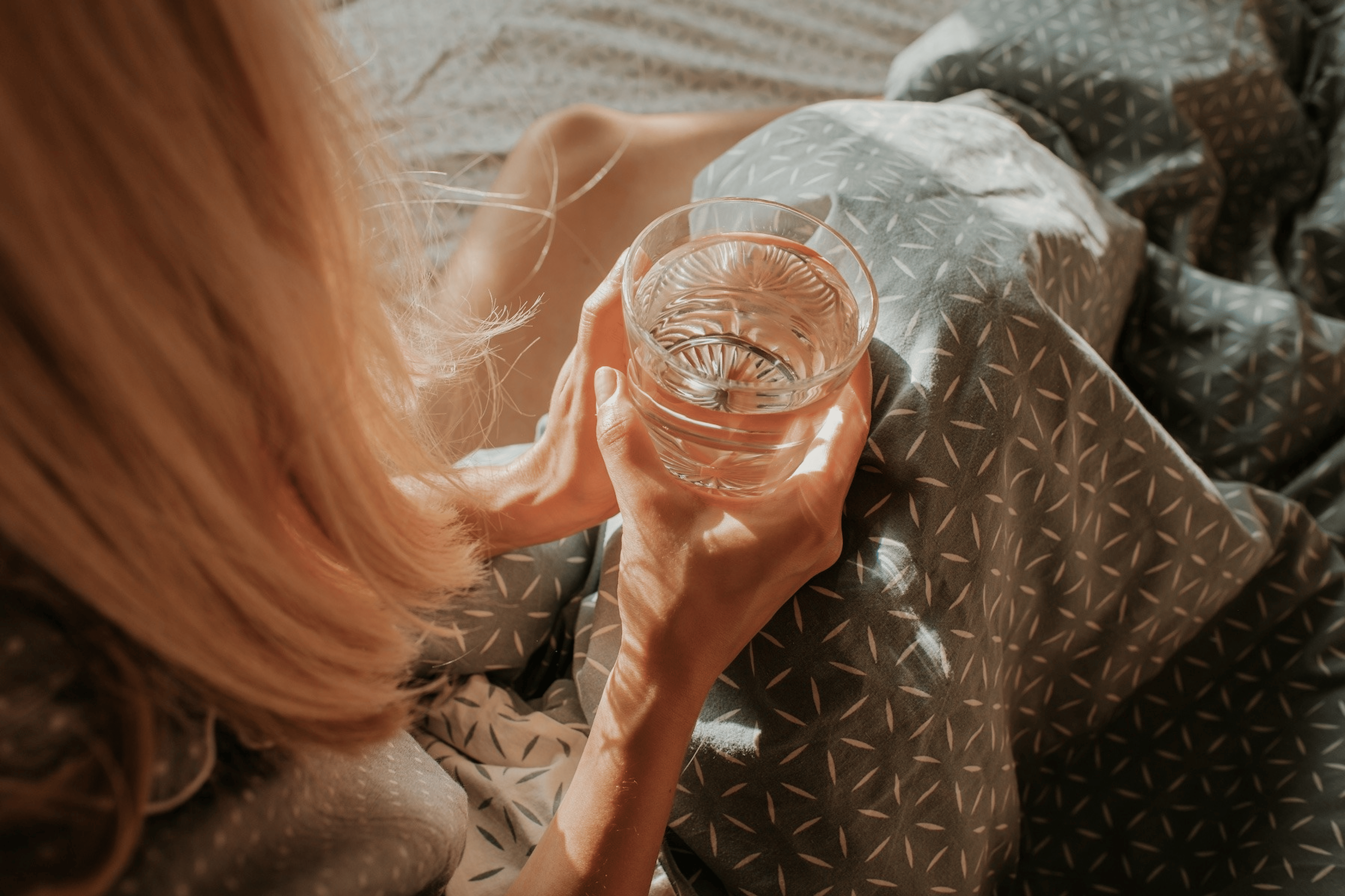 A close-up photo of a woman holding a glass of water in her hands as she sits in bed in the morning.