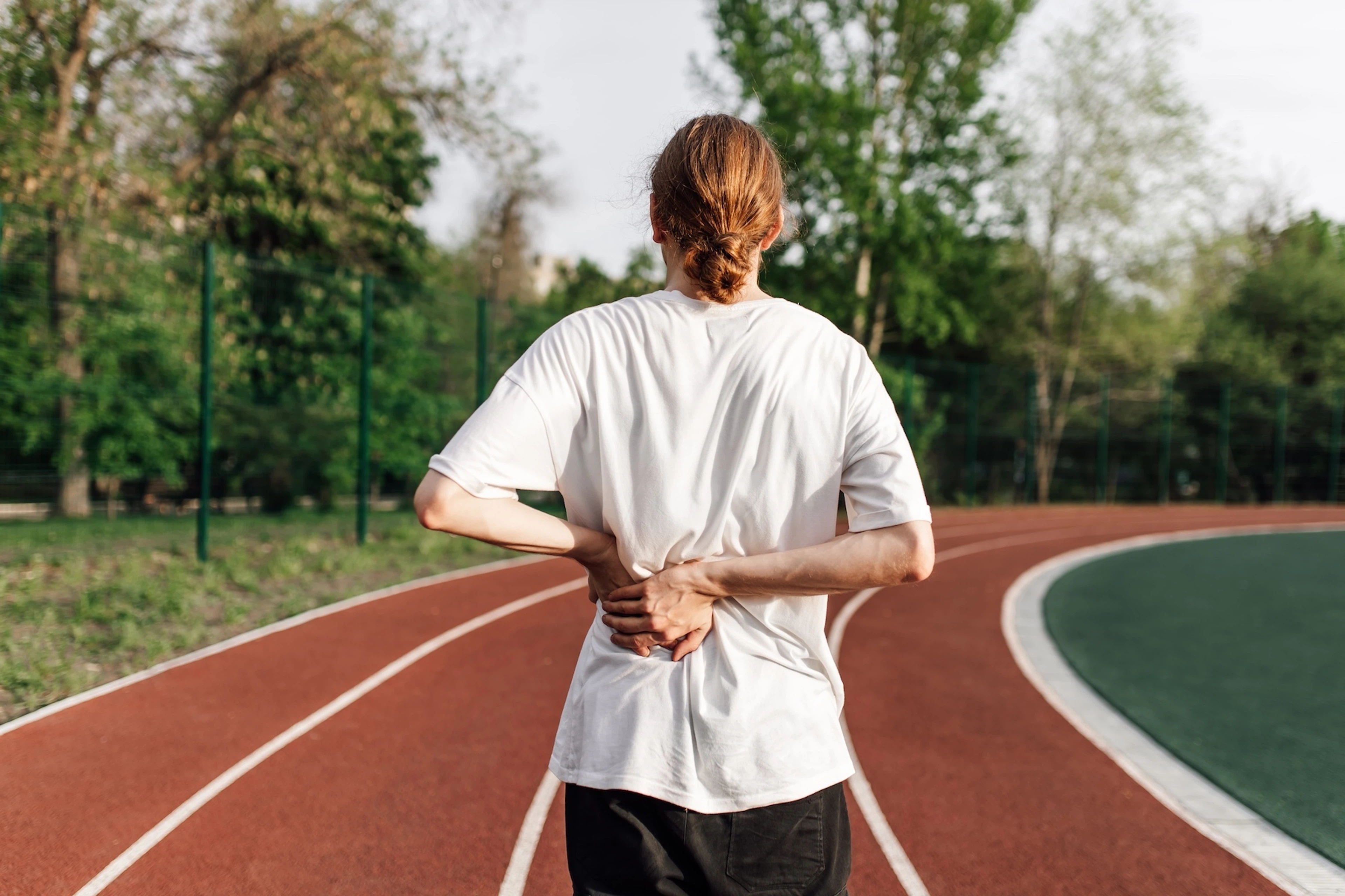 A photo of the back of an athlete who is on an outdoor track. They are feeling bloated after a workout and holding their back with their hands. 
