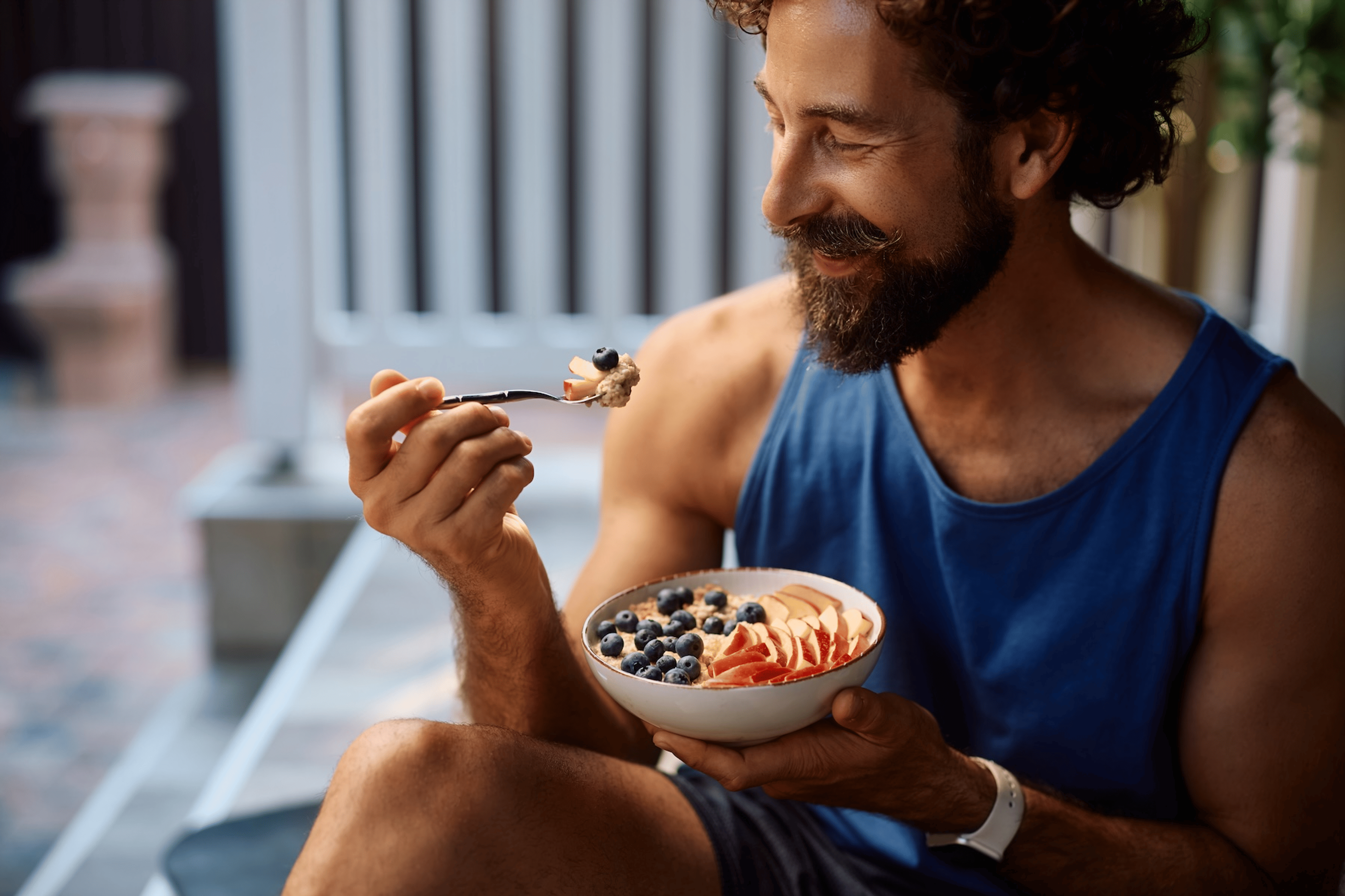 A man eating a bowl of oats topped with fresh fruit while sitting outside. He's eating carbs before his workout.