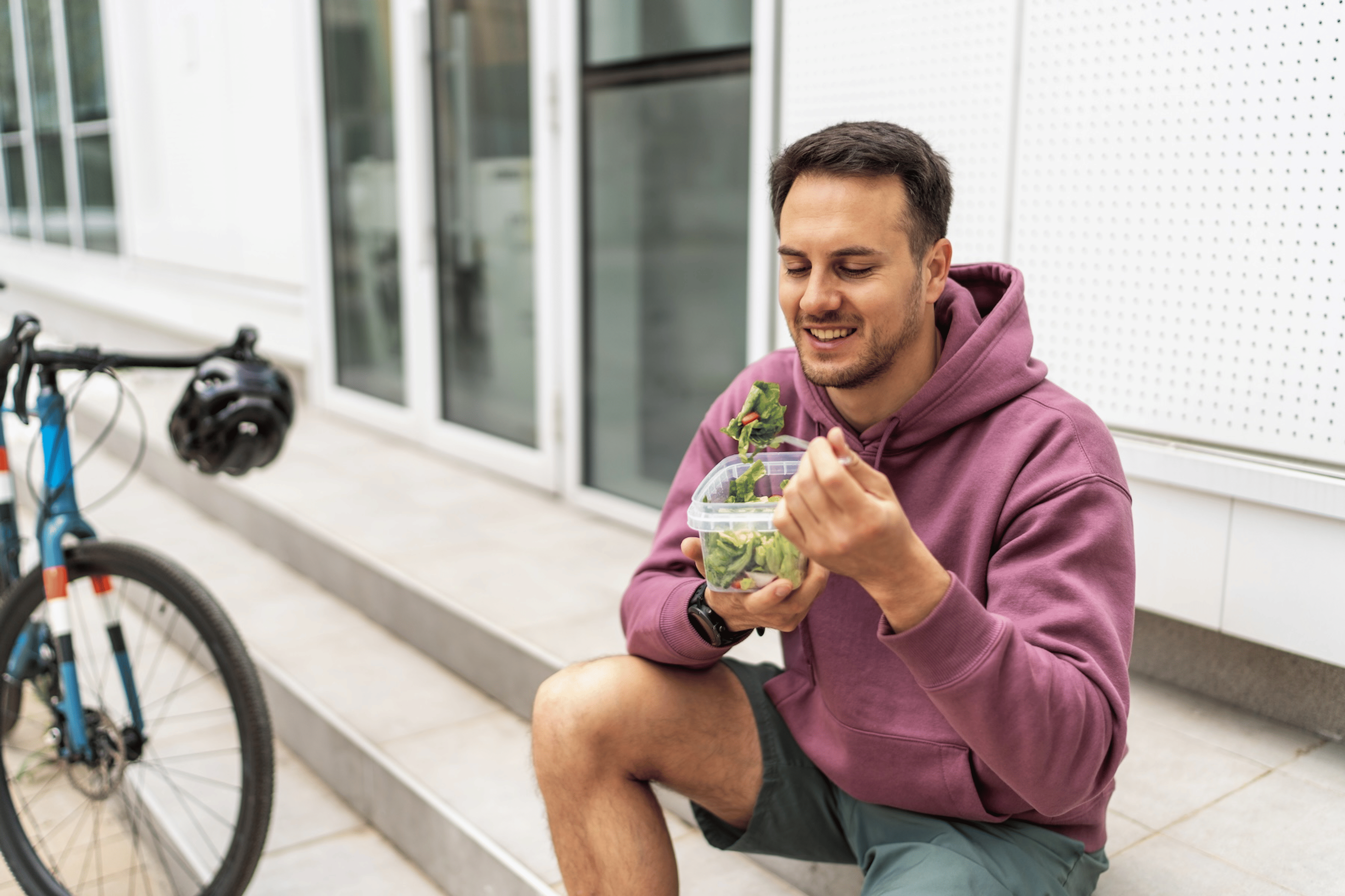 A man sitting on the steps outside near a bicycle eating a salad after his workout.
