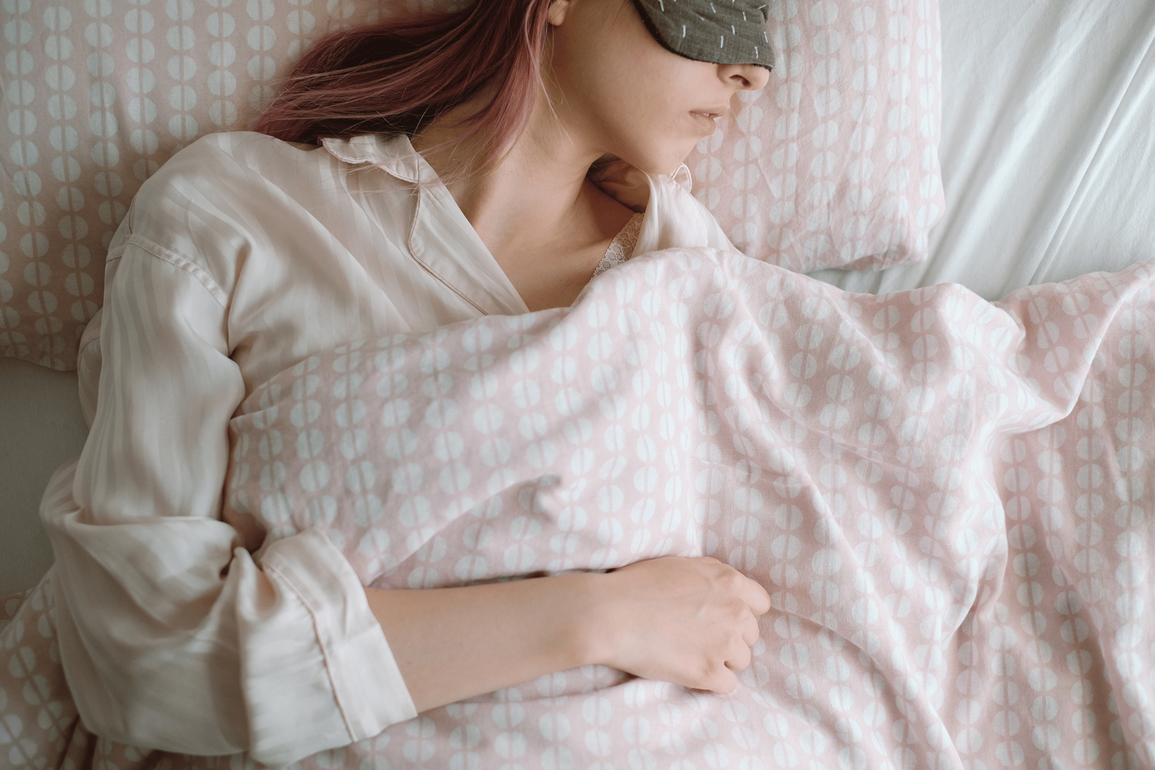 A woman with pink hair and an eye mask sleeping soundly in bed at the best temperature for sleep.