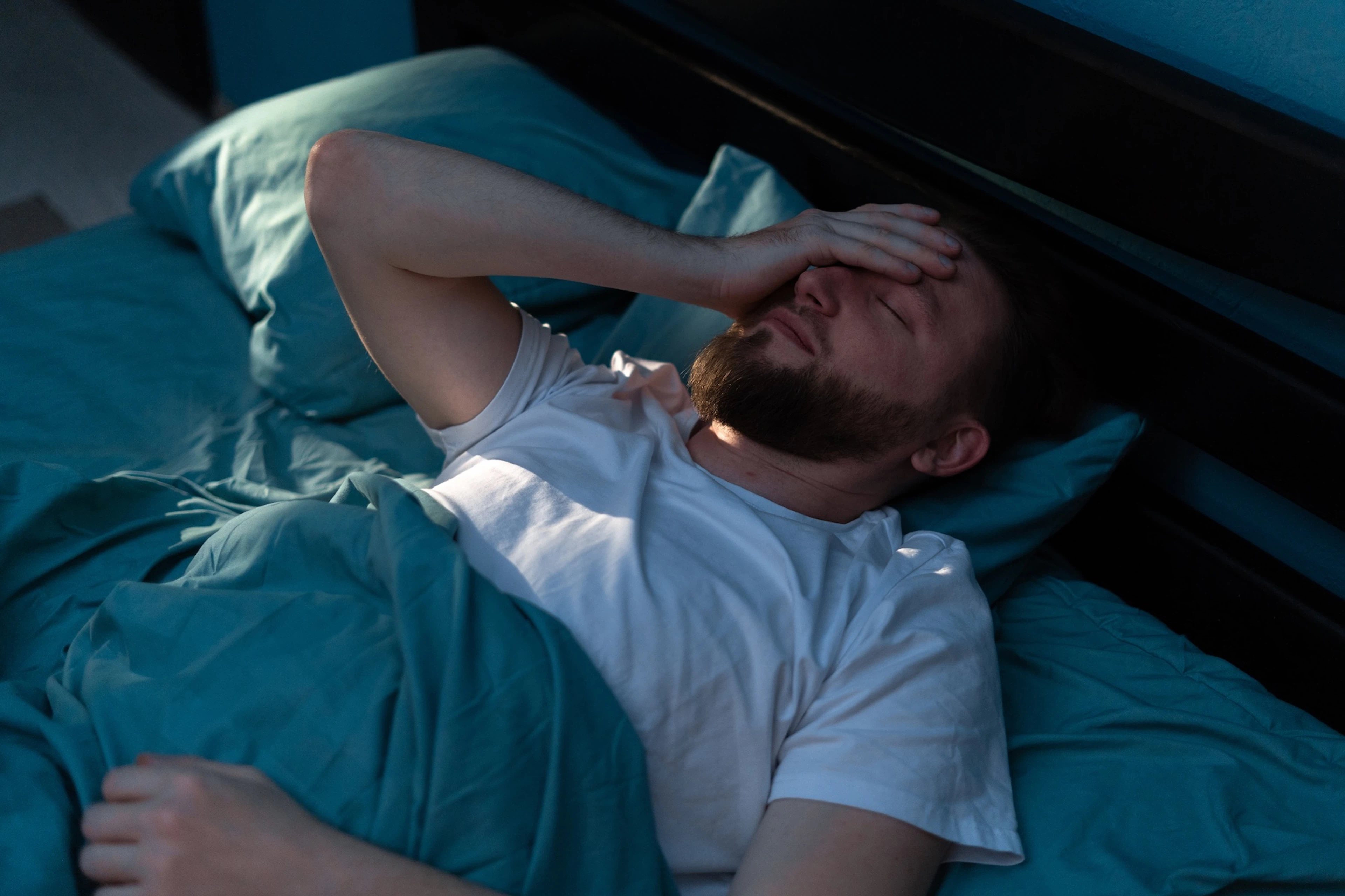 A man experiencing sleep anxiety. He's lying in bed at night with his hand wearily over his forehead.