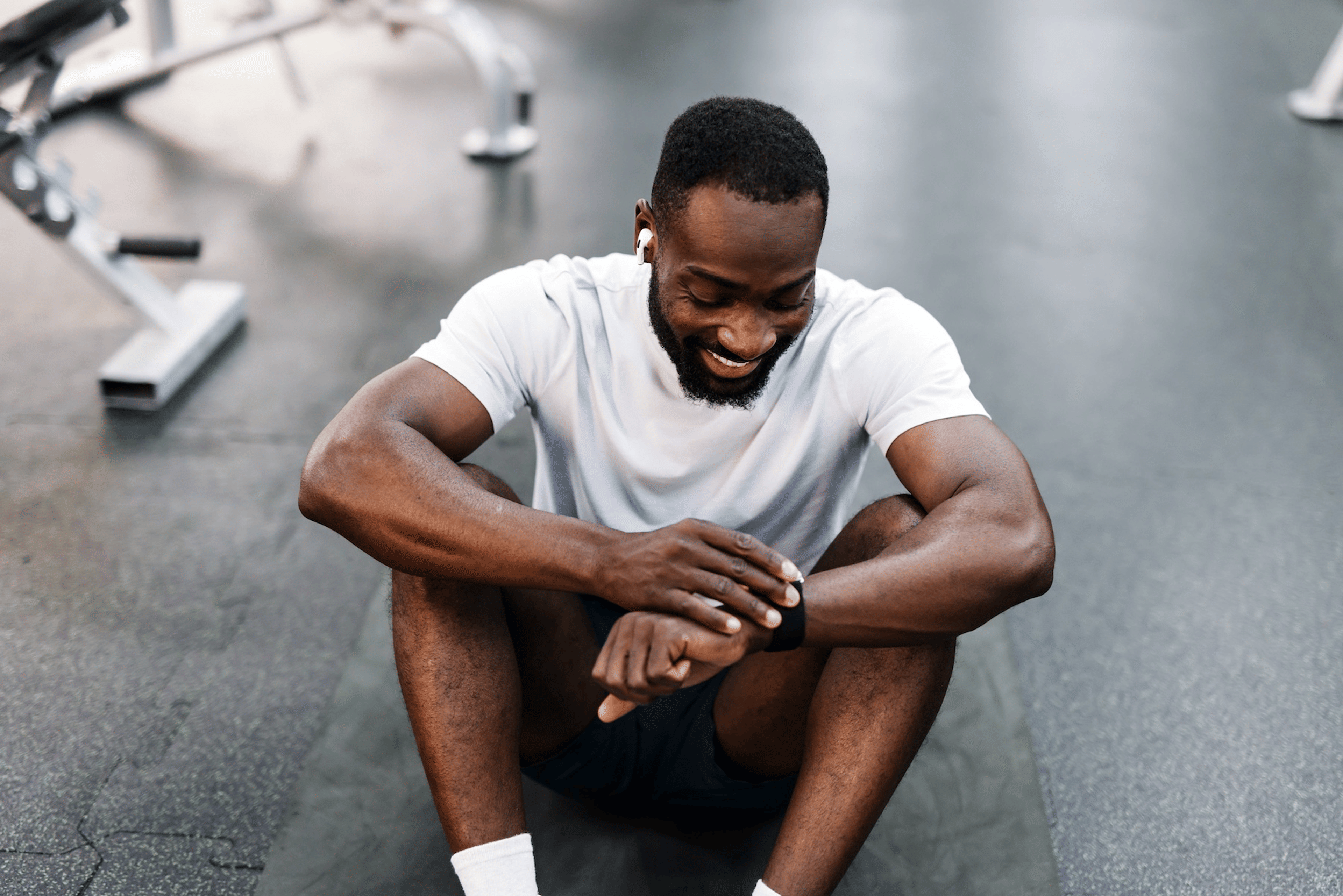A man sitting down at the gym, smiling and looking at his smart watch.