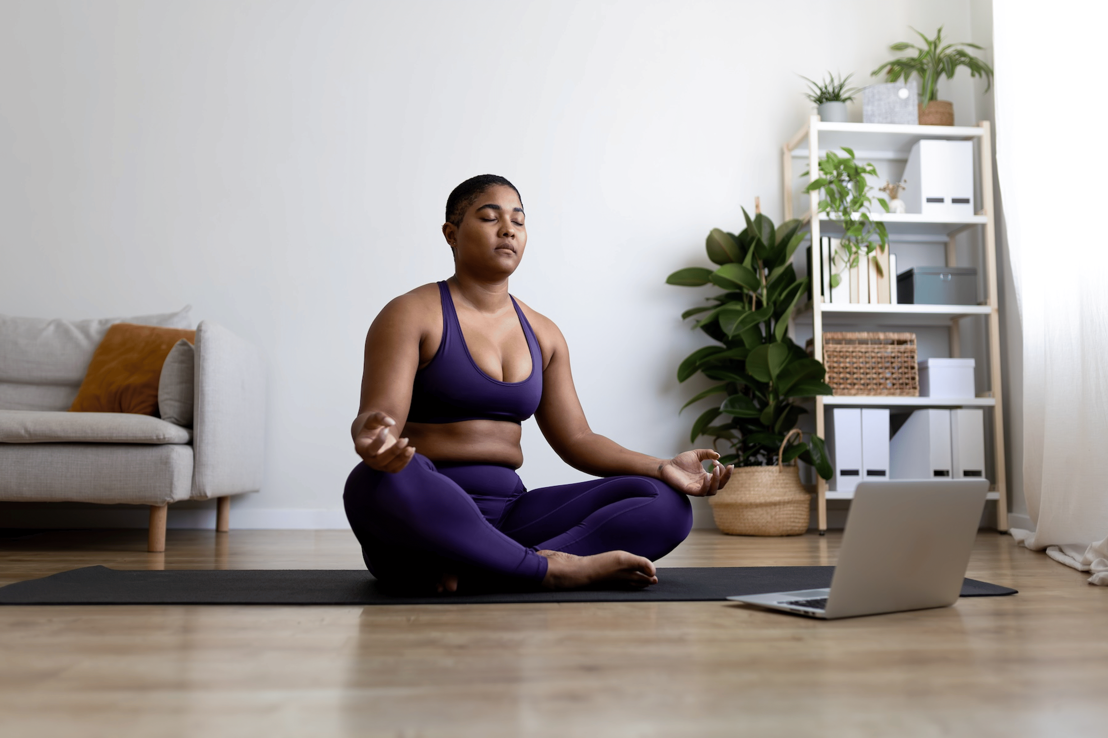 A woman meditating on a yoga mat in a cross-legged position. Her eyes are closed and her hands are resting on her knees. She has a laptop open in front of her that could be playing a guided meditation for self-confidence.