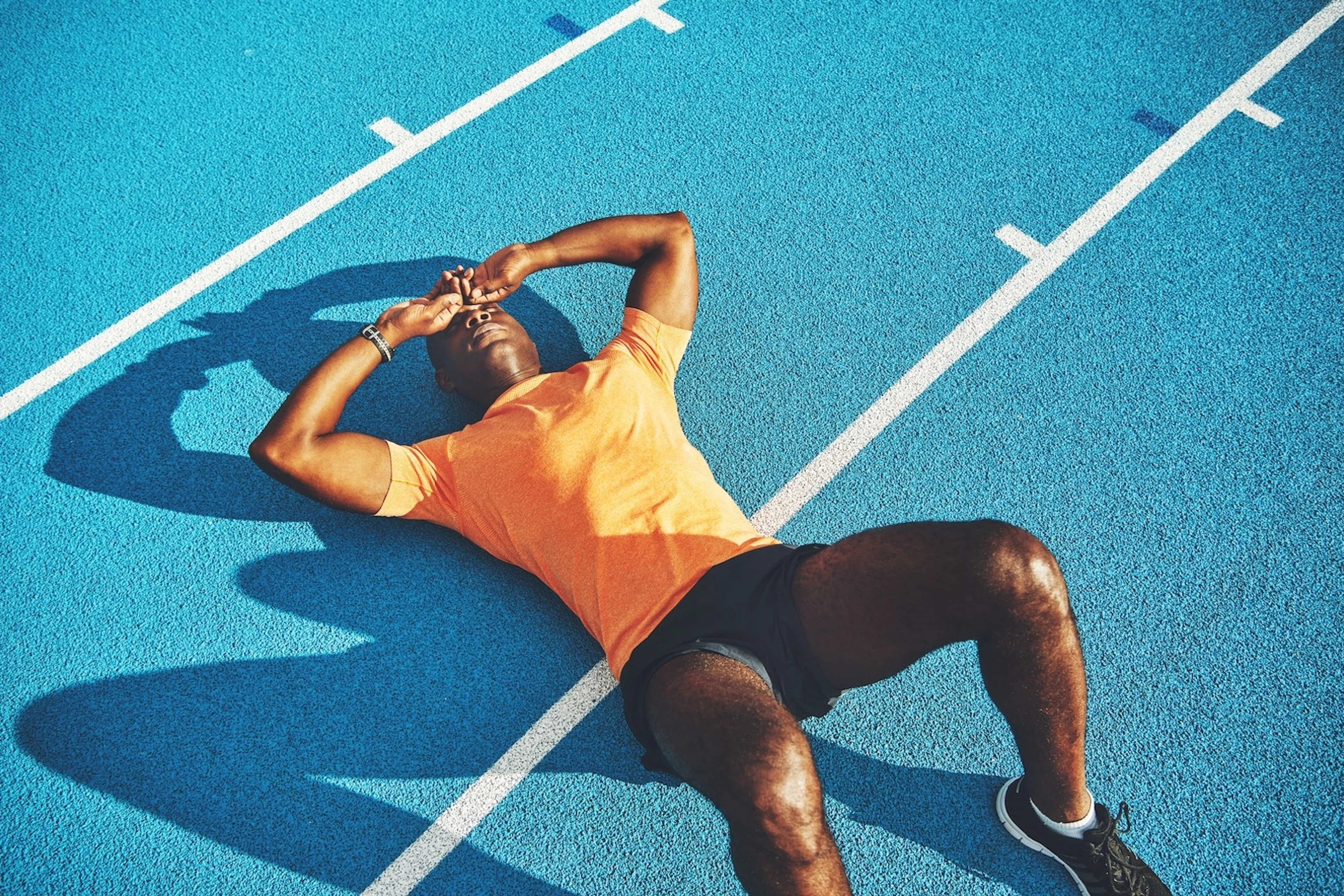 A male athlete experiencing bonking. He is exhausted, lying down on his back with his hands on his head, on a blue track.