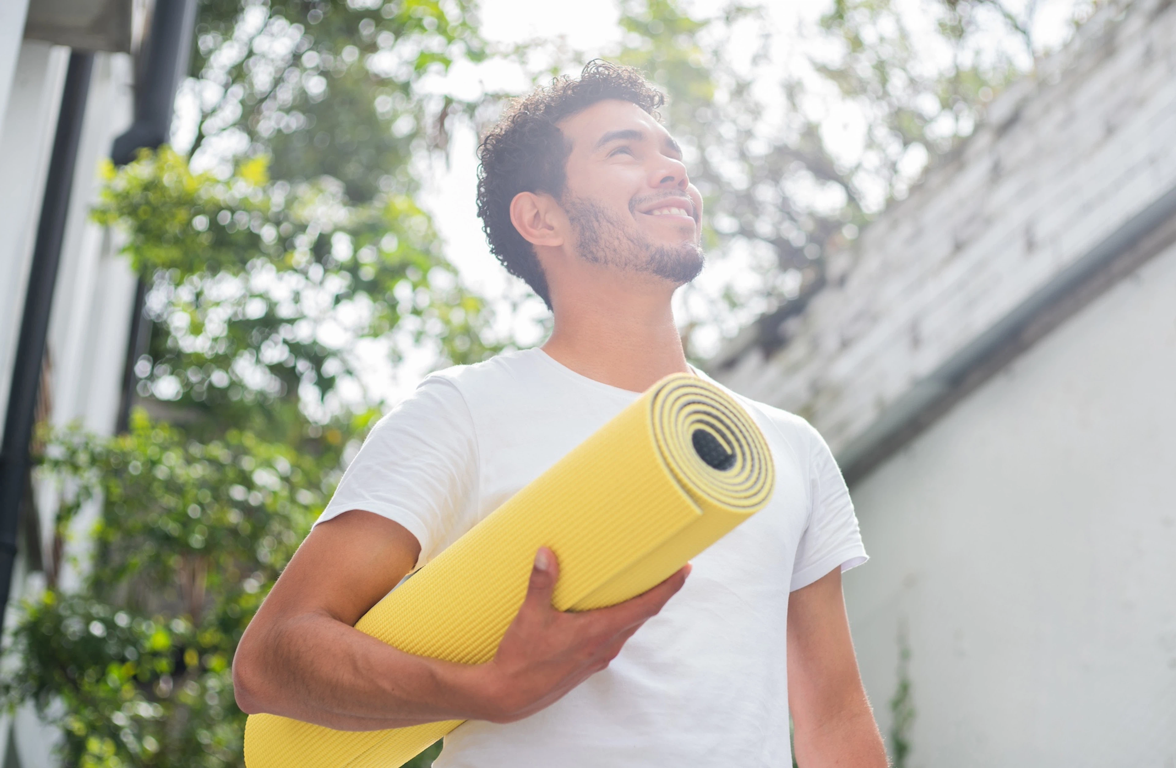 A man holding a yellow yoga mat and smiling while standing outside. Learn how to motivate yourself to work out here.