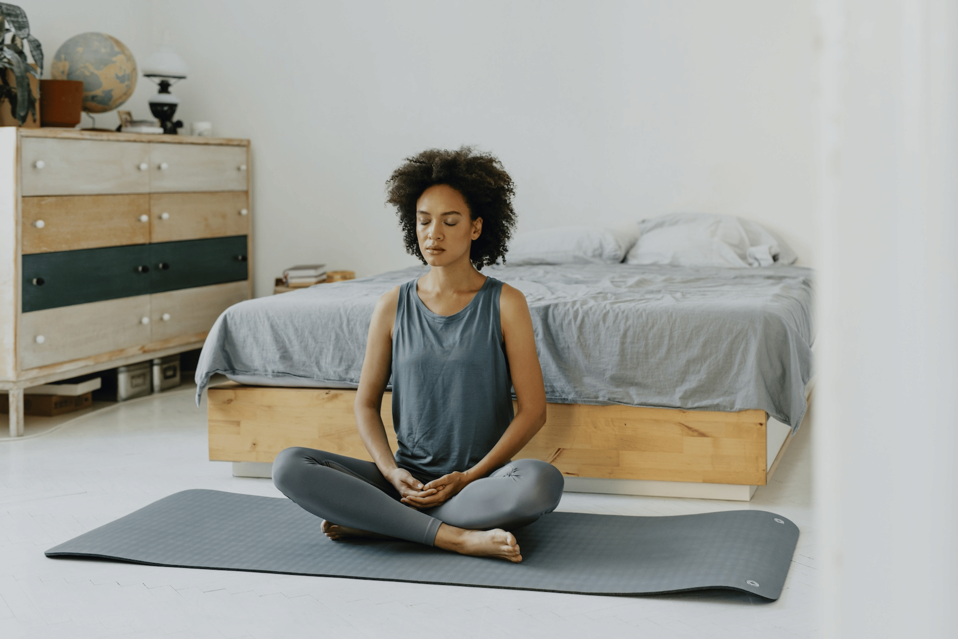 A woman practicing a loving-kindness meditation in her bedroom while sitting criss-cross on a yoga mat.
