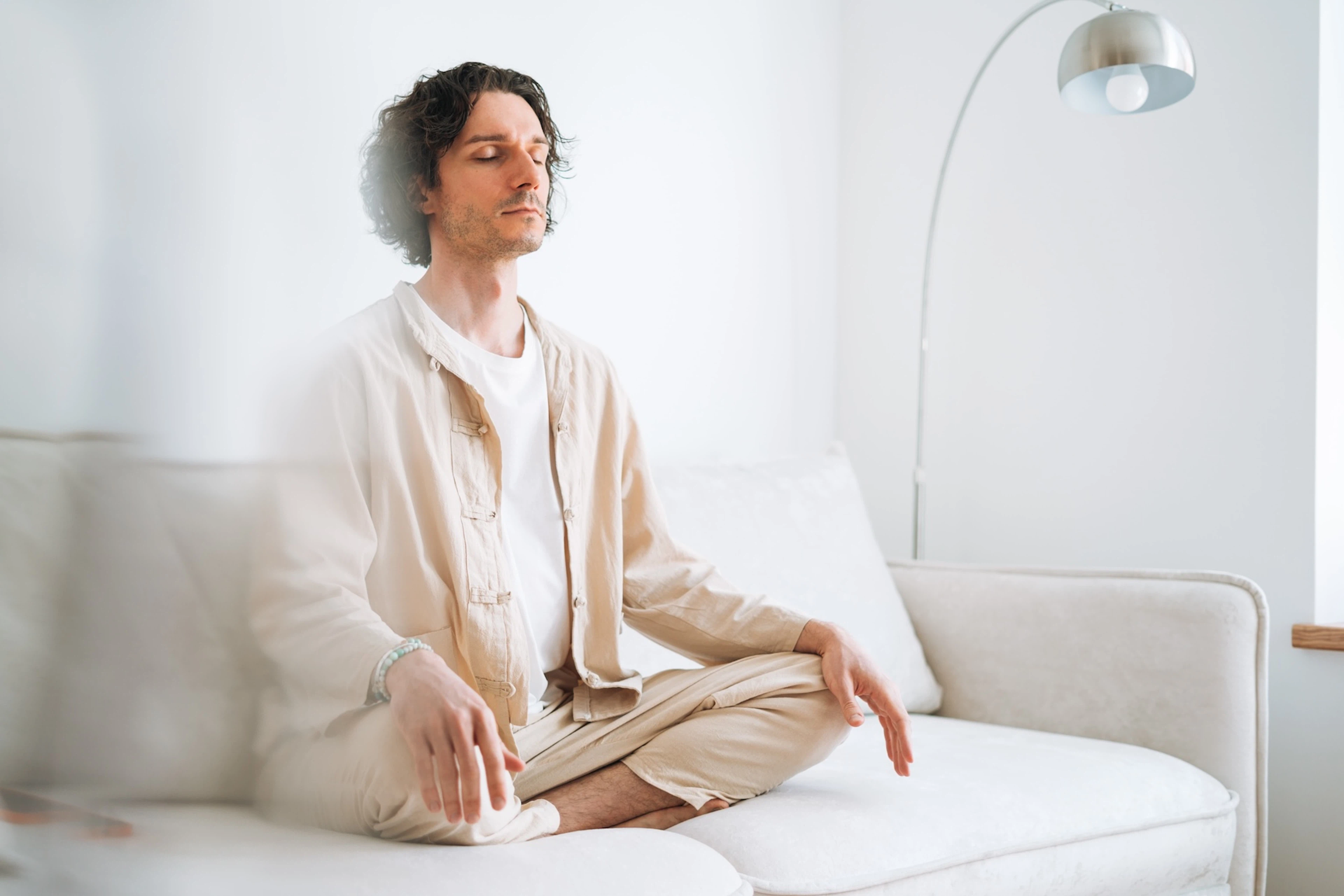 A young man sitting cross-legged on a white couch in a bright room and meditating.