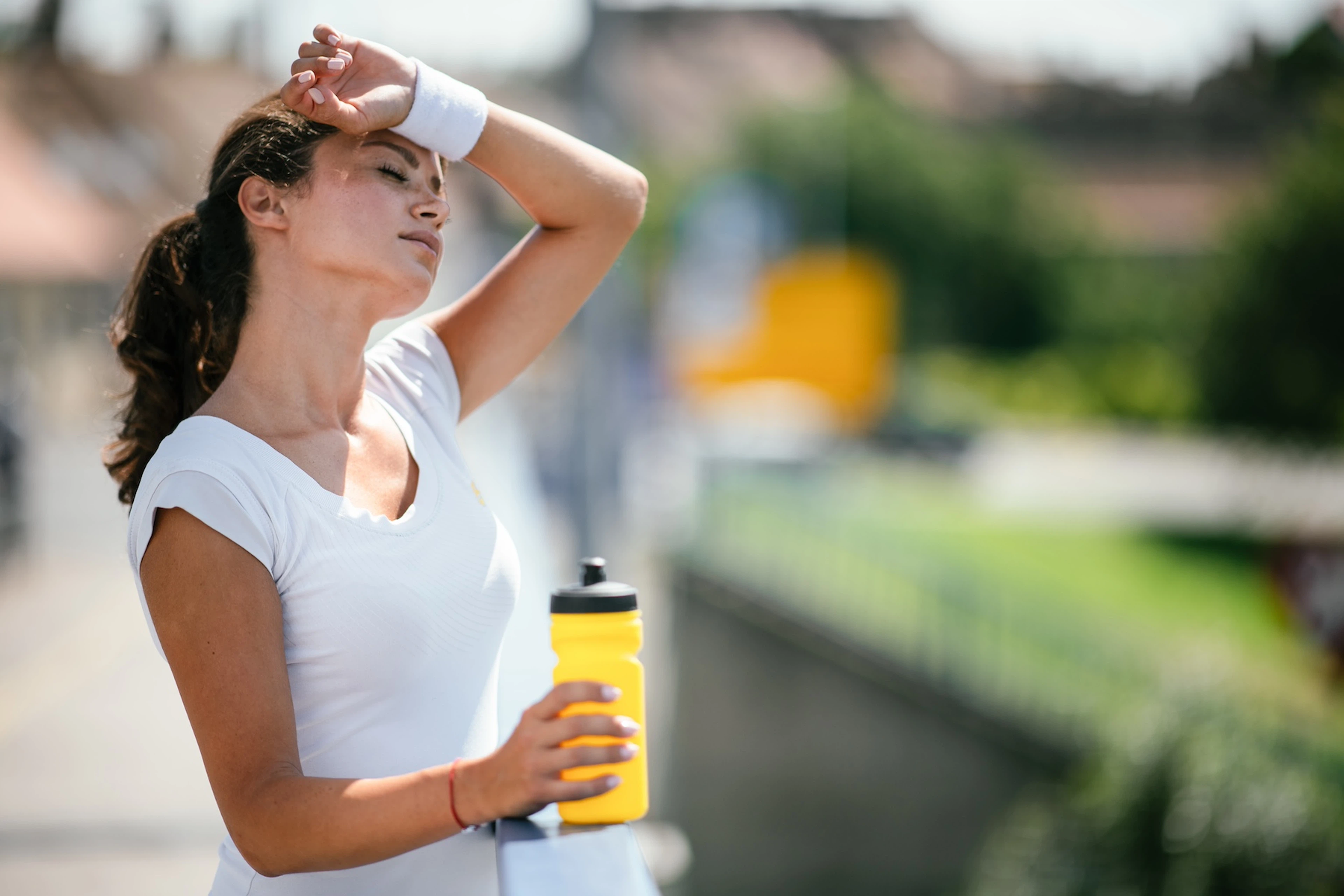 A tired athlete rubbing sweat off her forehead with her sweatband and drinking from a yellow water bottle. Learn how to rehydrate in this article.