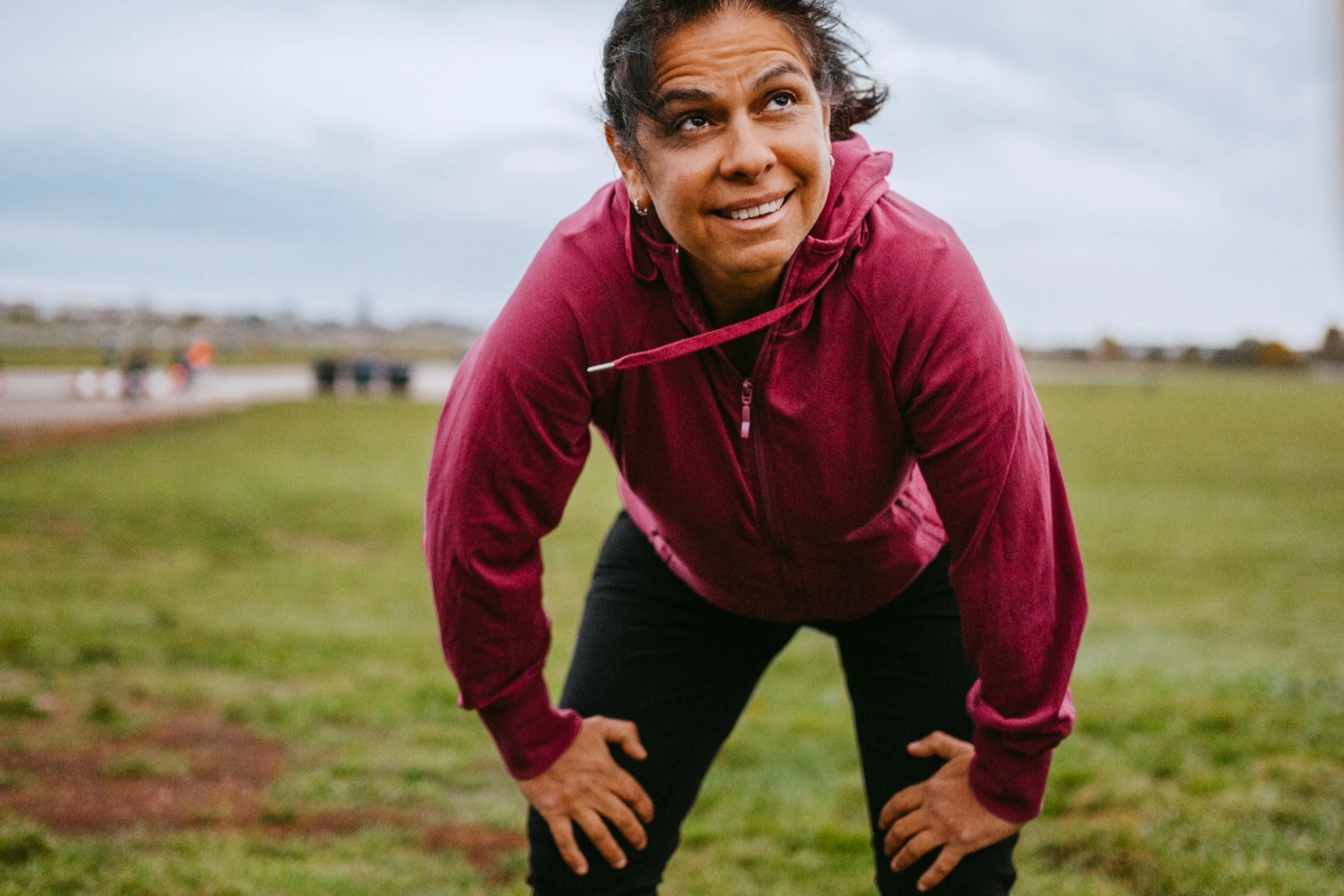 A happy, tired woman putting her hands on her knees as she takes a breather during an outdoor workout.