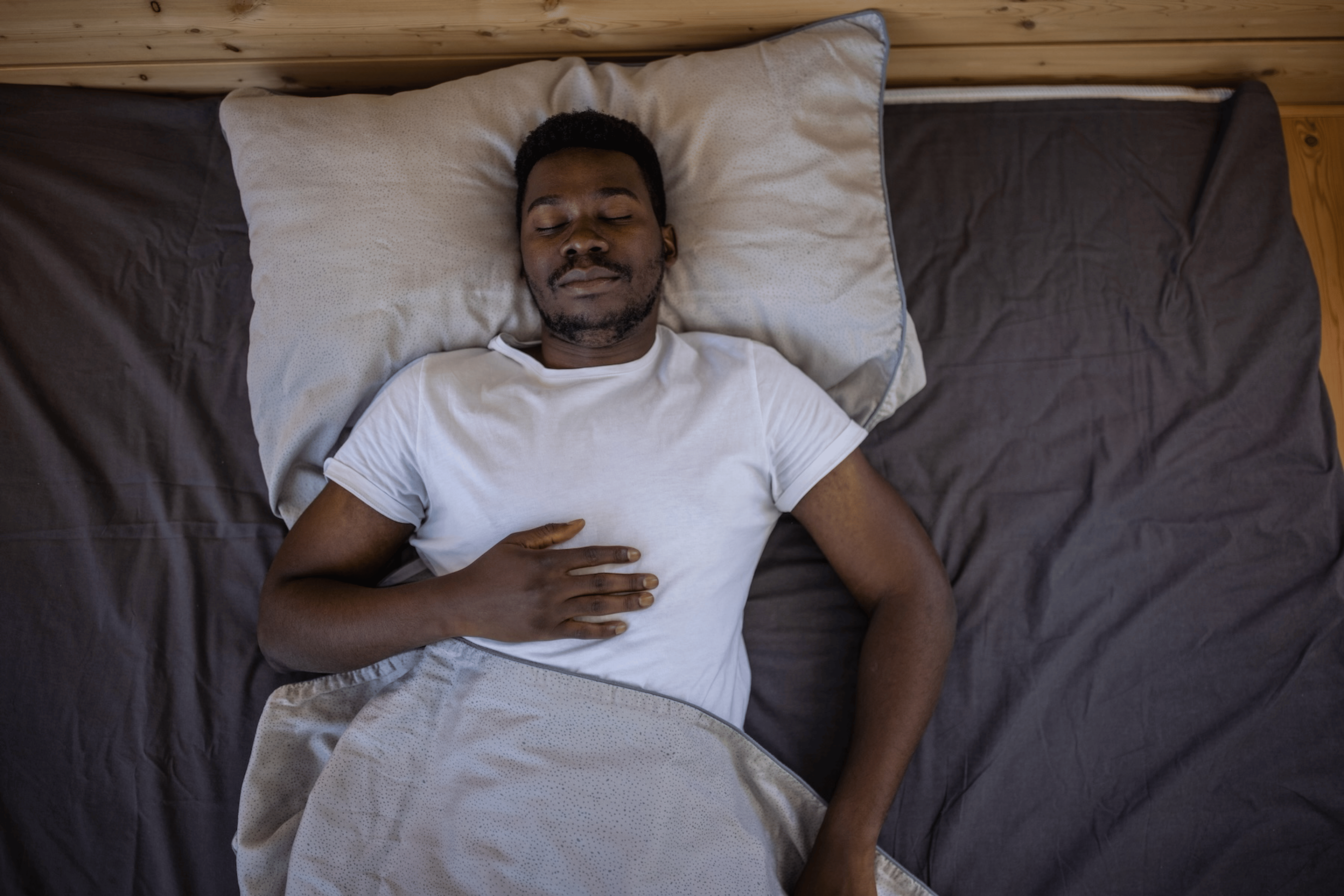 A man practicing breathing exercises for sleep. He's lying on his back in bed, eyes closed,  with his right hand on his upper abdomen.