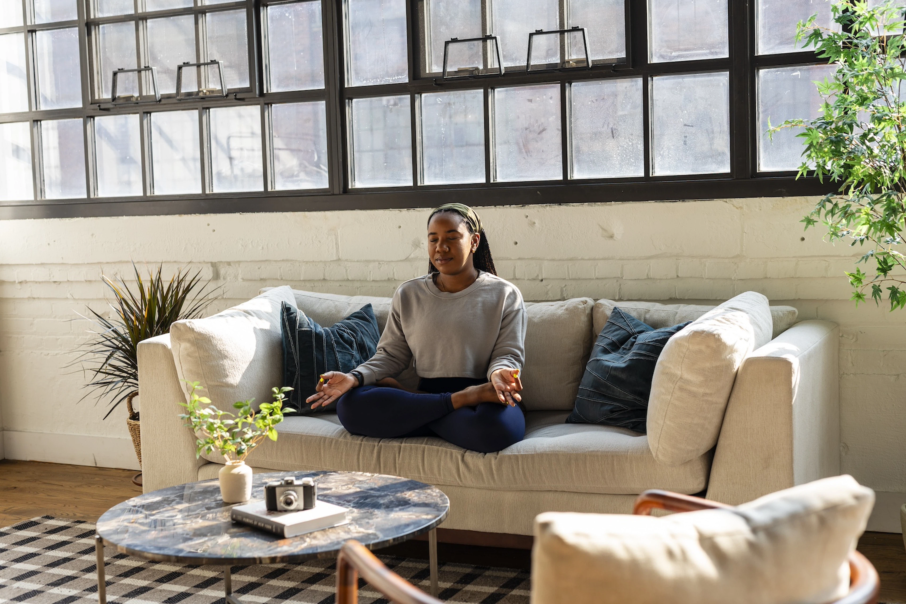 A woman practicing healing meditation. She's sitting in Lotus Pose on a couch at home in a sunny living room.
