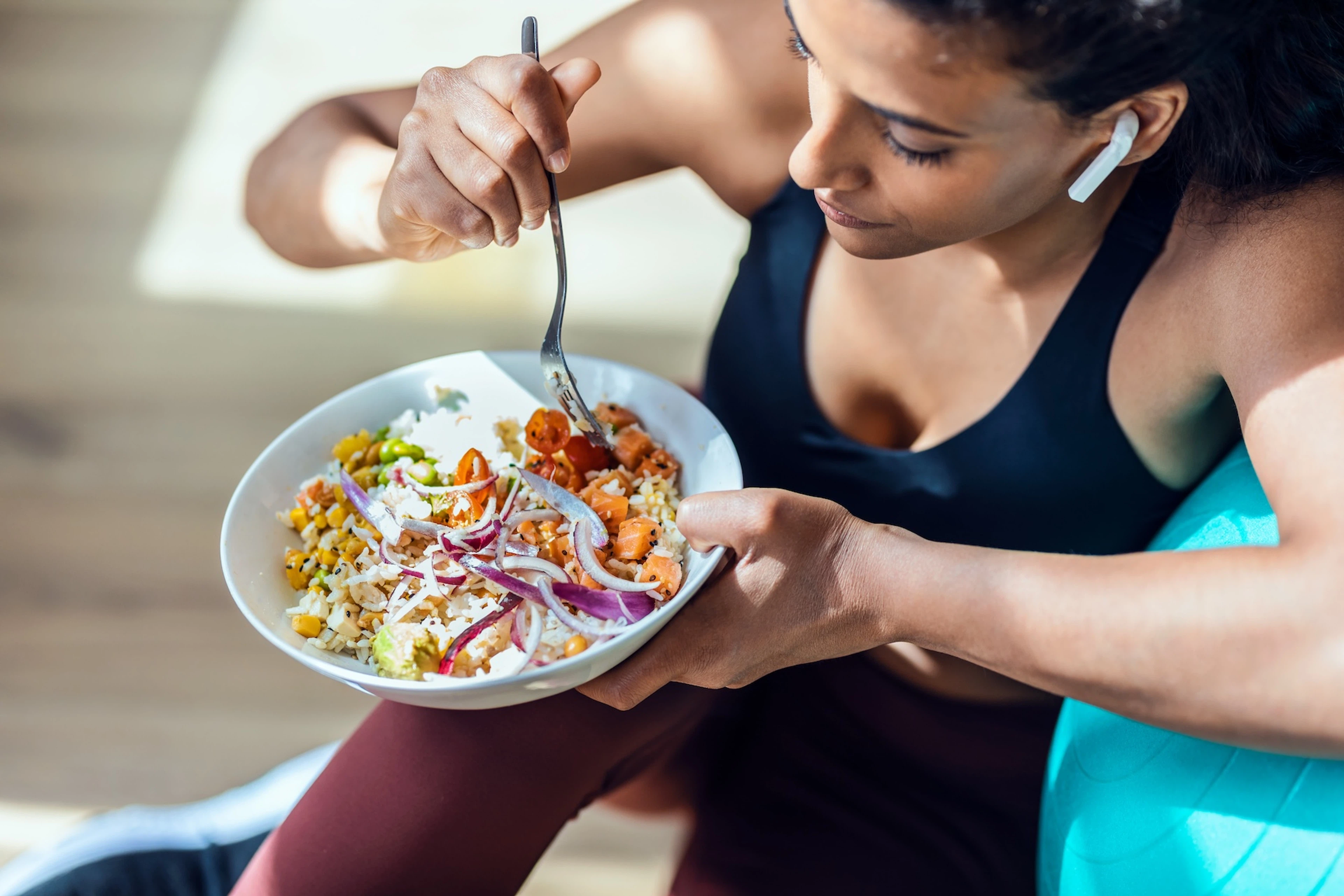 A woman eating dinner (a veggie-packed grain bowl) before or after a workout. She's in workout clothes and is leaning against a big exercise ball. 