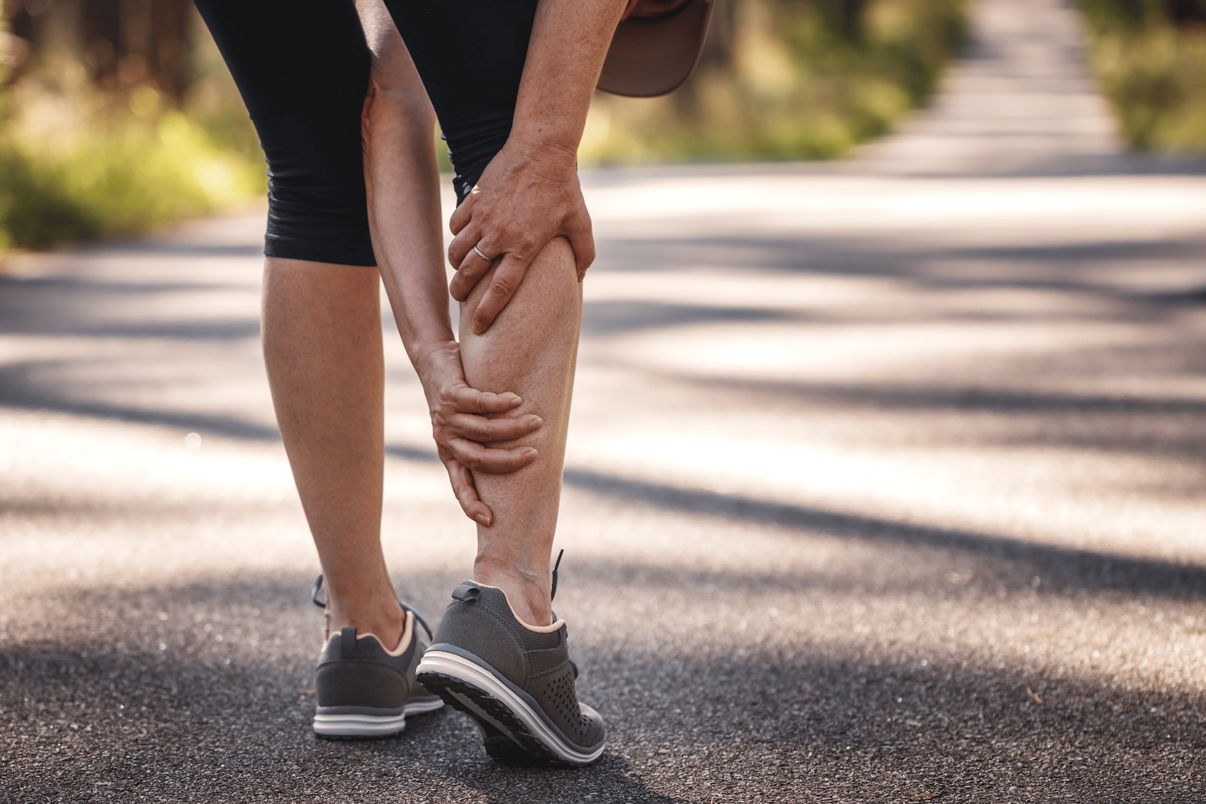 A close-up photo of a woman grabbing her cramping calf muscle during an outdoor run.