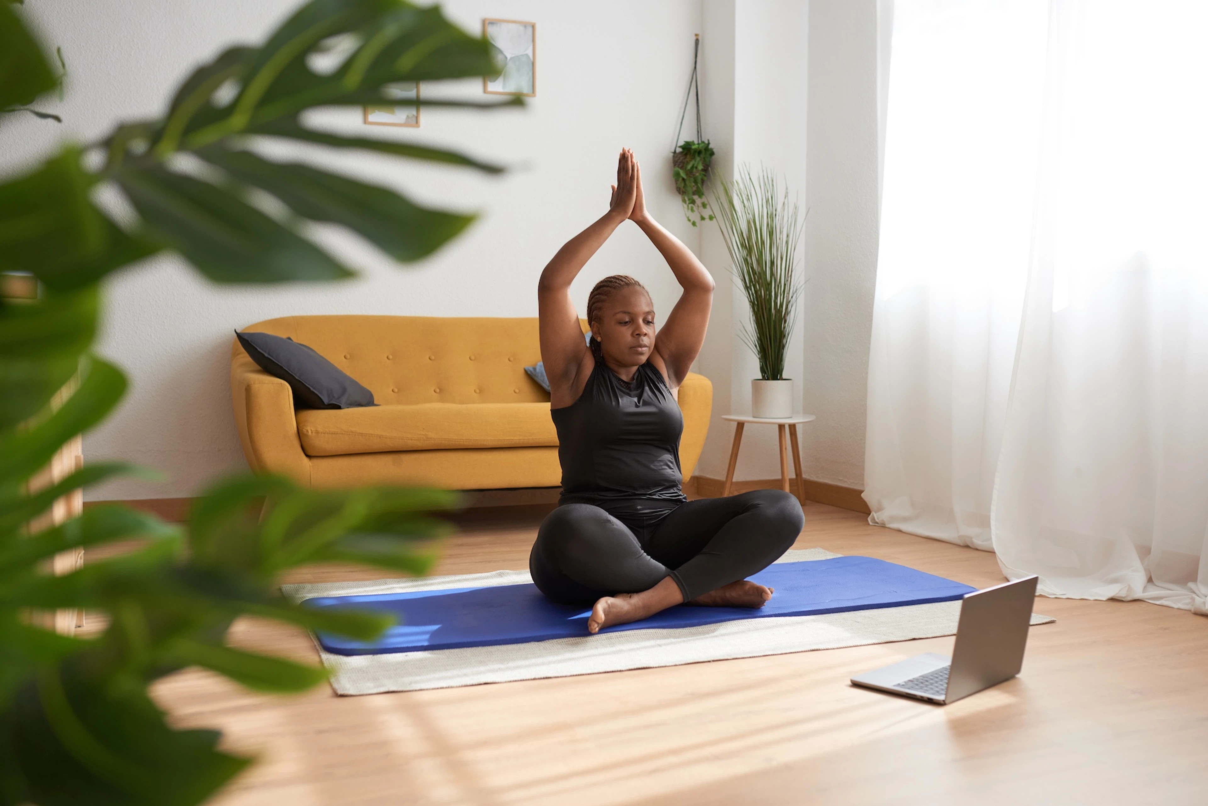A woman doing guided yoga on a mat at home. She's sitting cross-legged with her hands joined together above her head.
