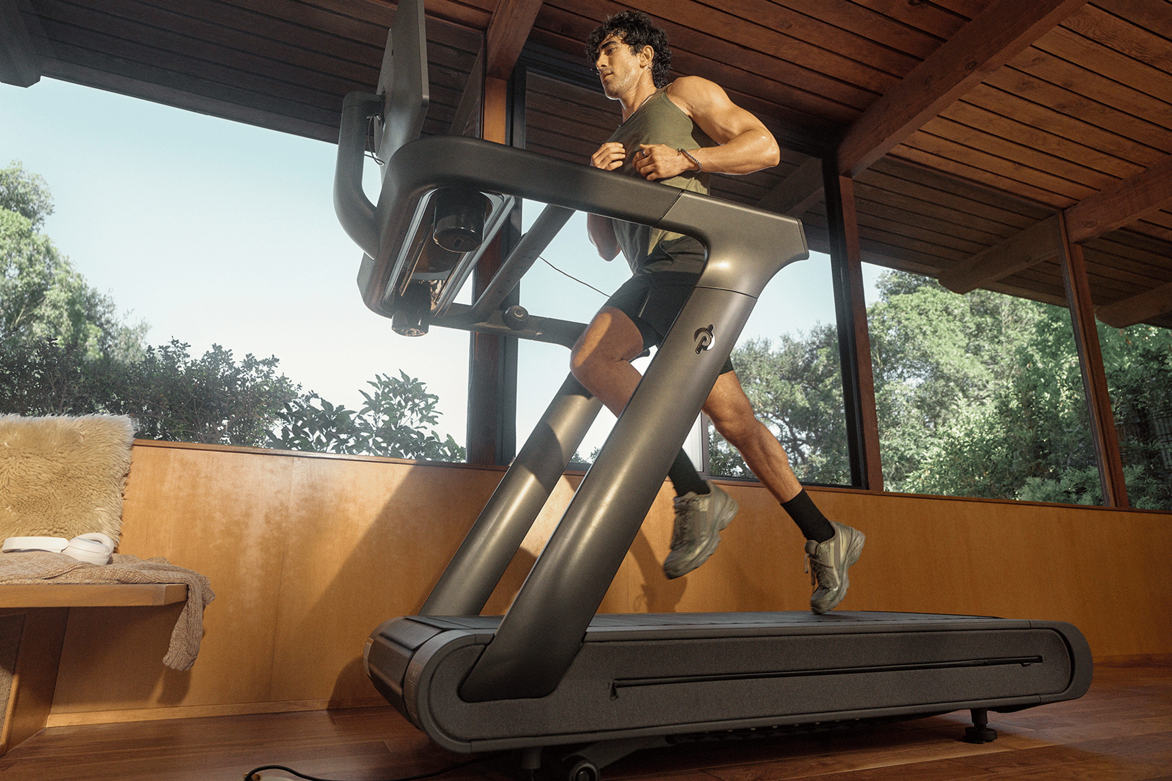 Man running on a Peloton Tread treadmill inside a room with large glass windows and wooden ceiling.