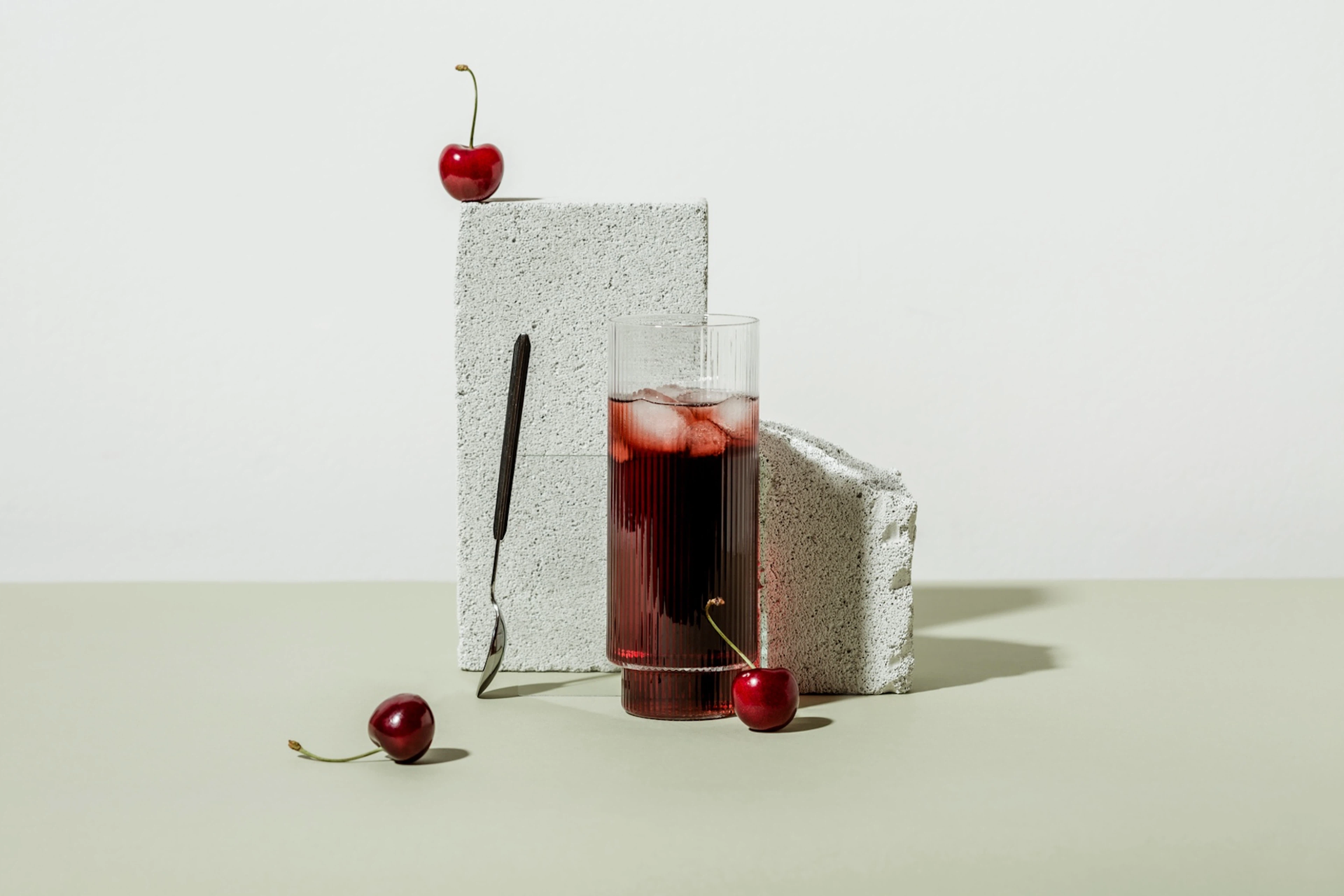 A glass of tart cherry juice propped against a cinder block with tart cherries and a spoon nearby.