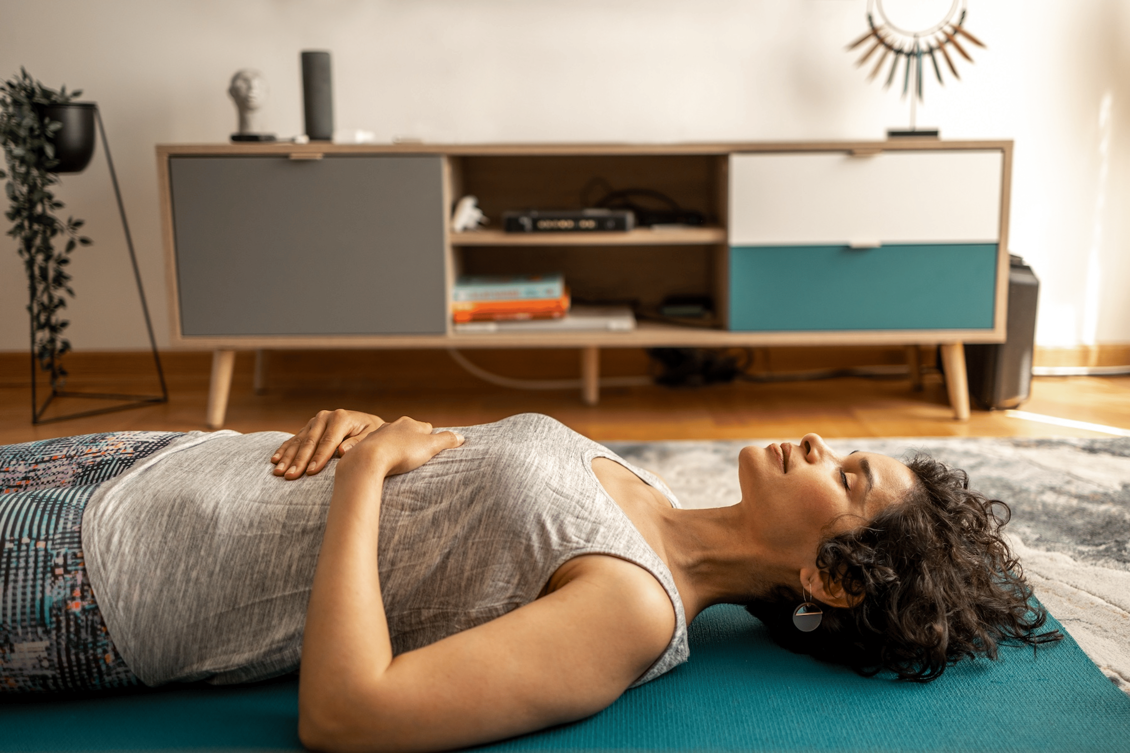 A woman lying on her back on a yoga mat at home, meditating. Her eyes are closed and her hands are resting on her belly.