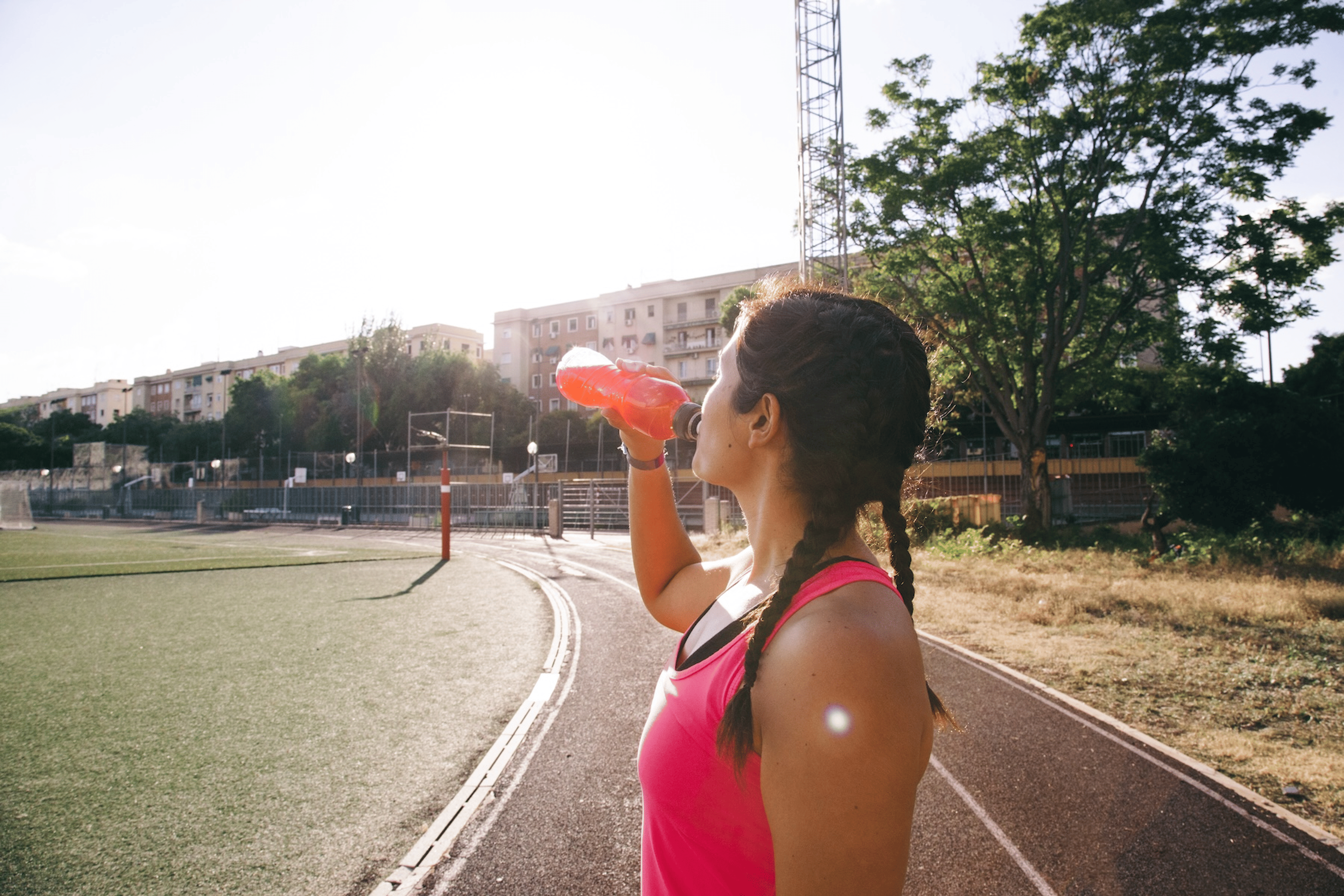 A side view of a woman drinking an orange-red electrolyte drink on an outdoor track.