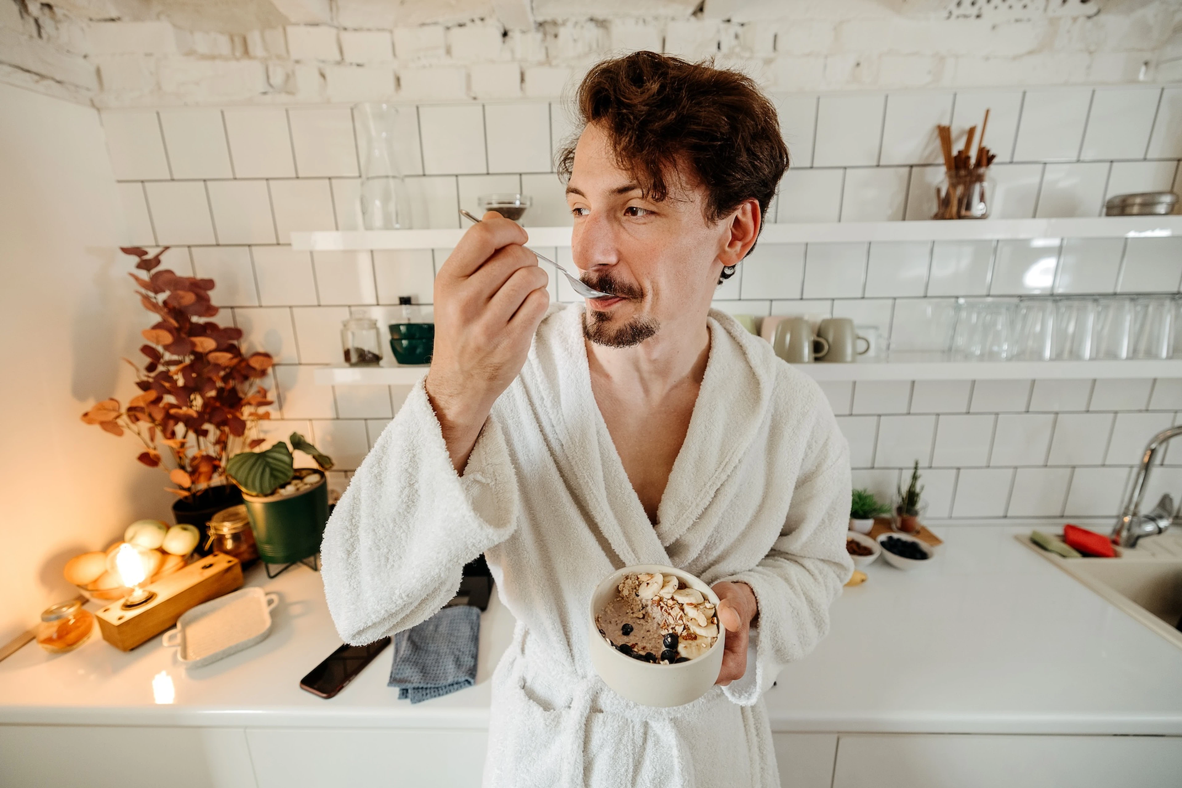 A man in his bathrobe eating oatmeal and fruit in his kitchen.