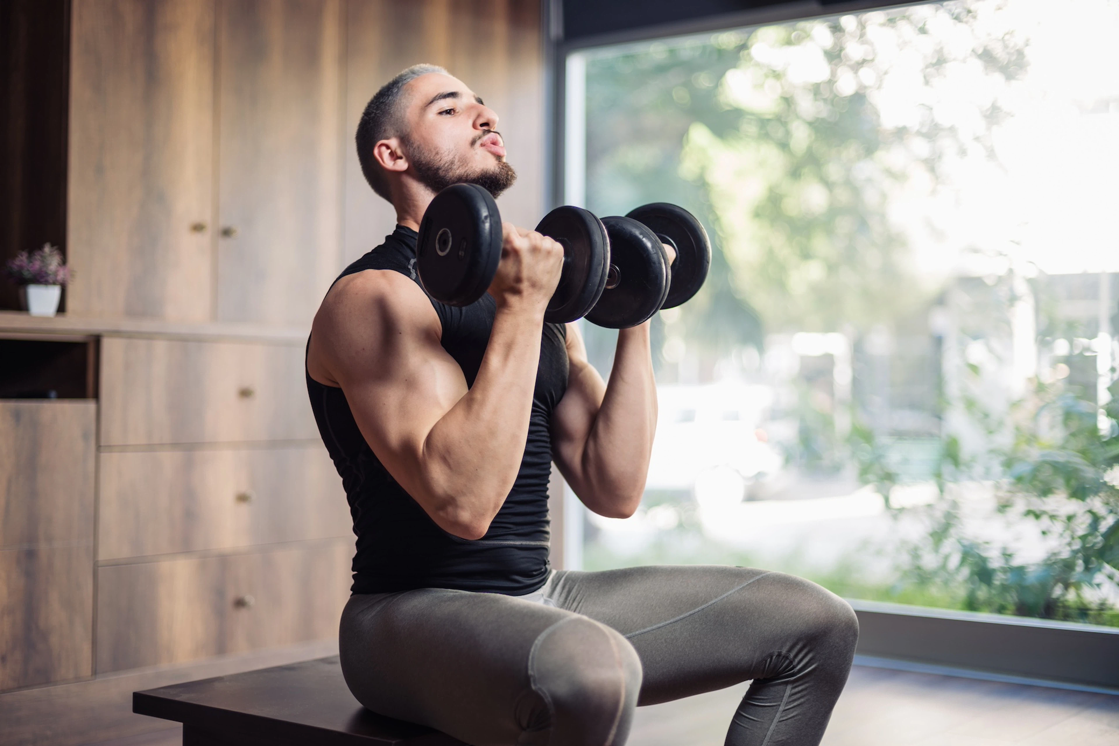 A man sitting down and lifting dumbbells in a sunny, airy gym.