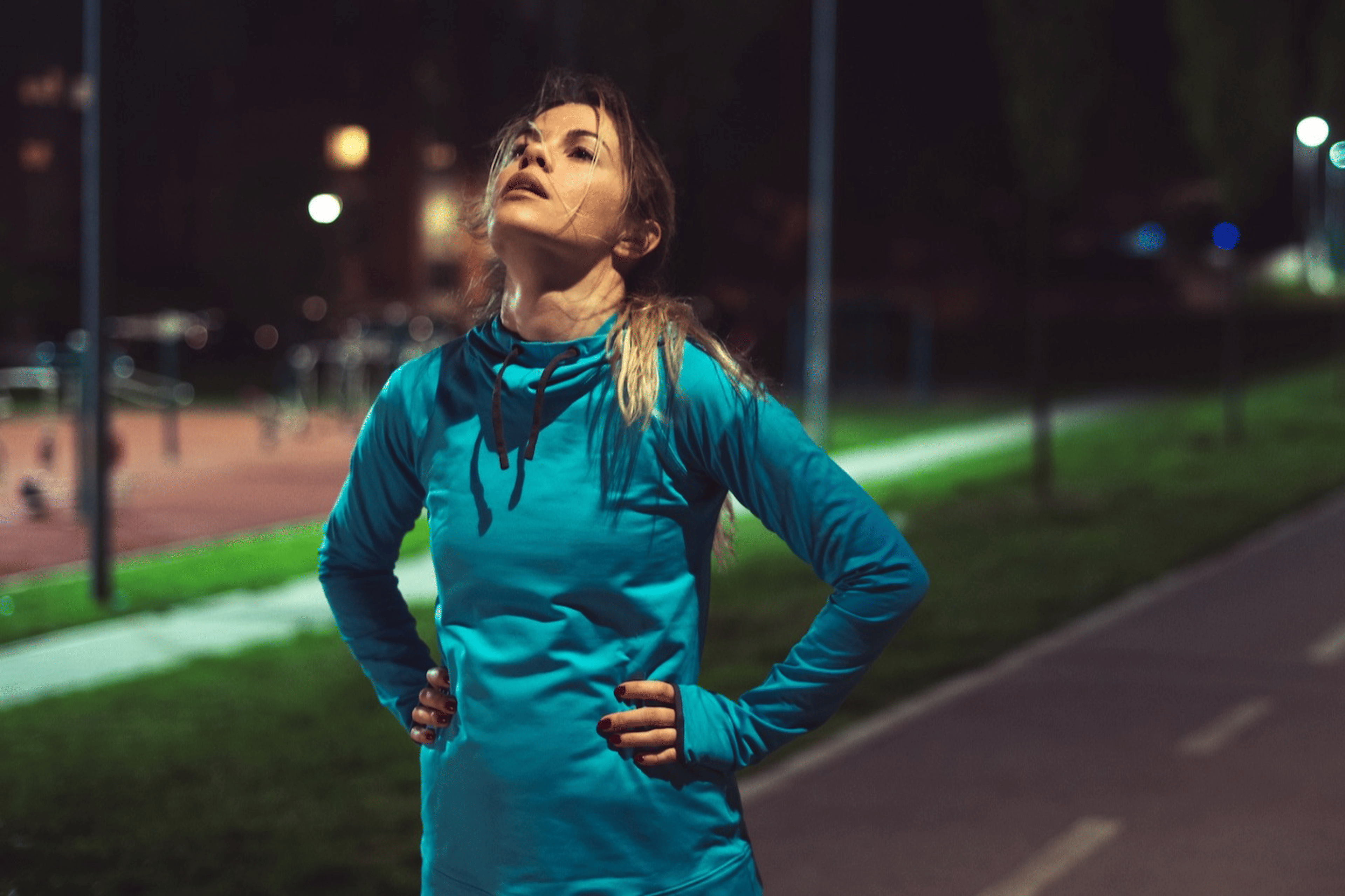 An exasperated athlete on an outdoor running path in the evening, with her hands on her hips and head pointing up to the sky. She has lost motivation to work out.