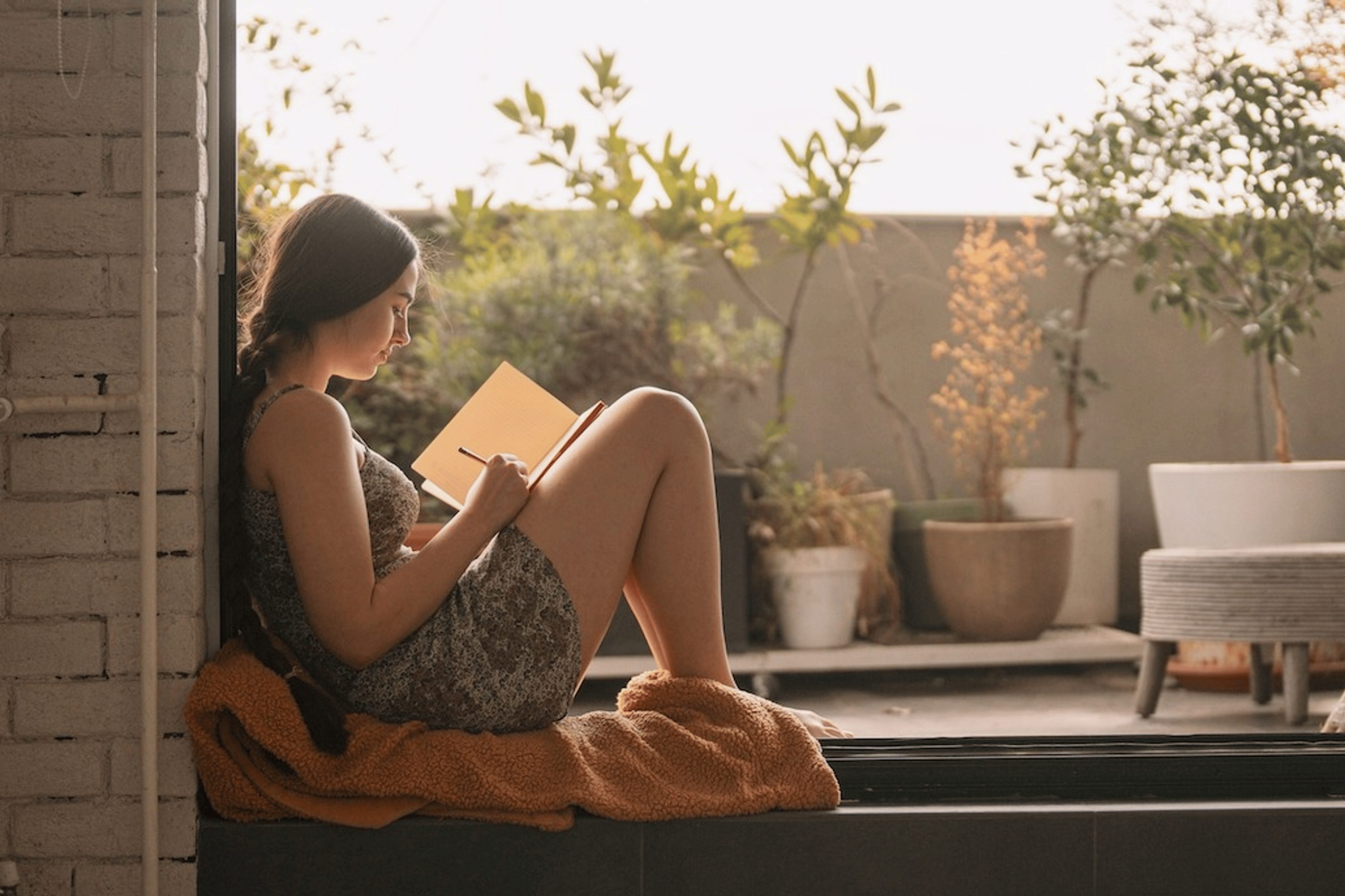 A woman writing affirmations for self-esteem in her journal. She's sitting on her apartment terrace on a nice day, leaning against the open doorway.