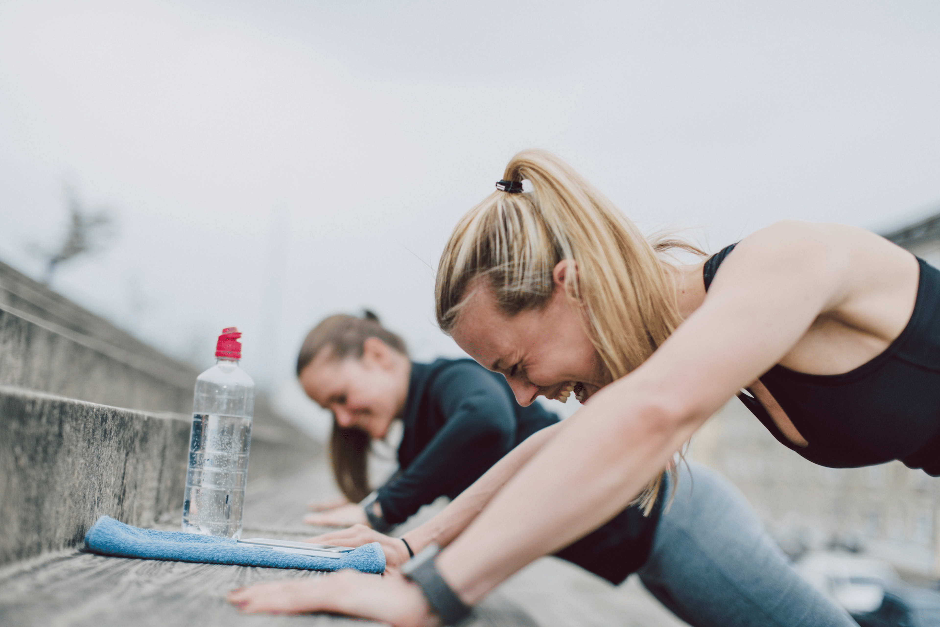 Two women doing push-ups together against an outdoor staircase and laughing. They're engaging in friendly competition.