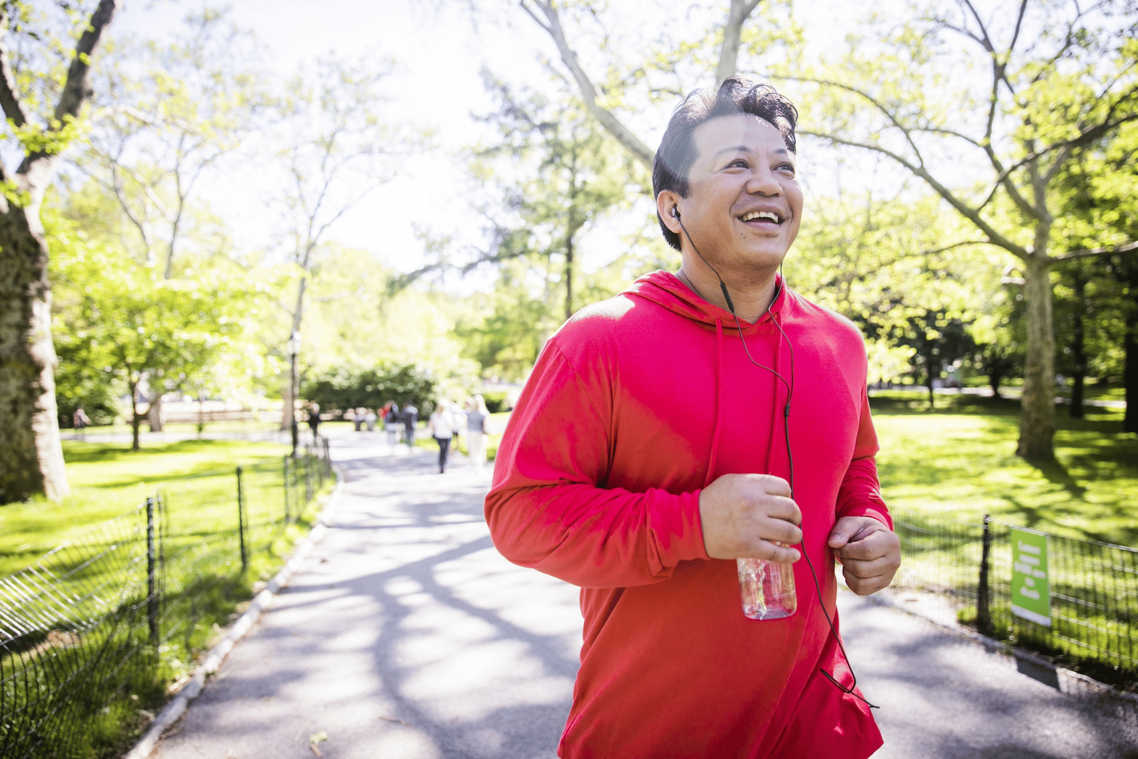 An adult man happily running through the park. He's listening to wired headphones and holding a water bottle.