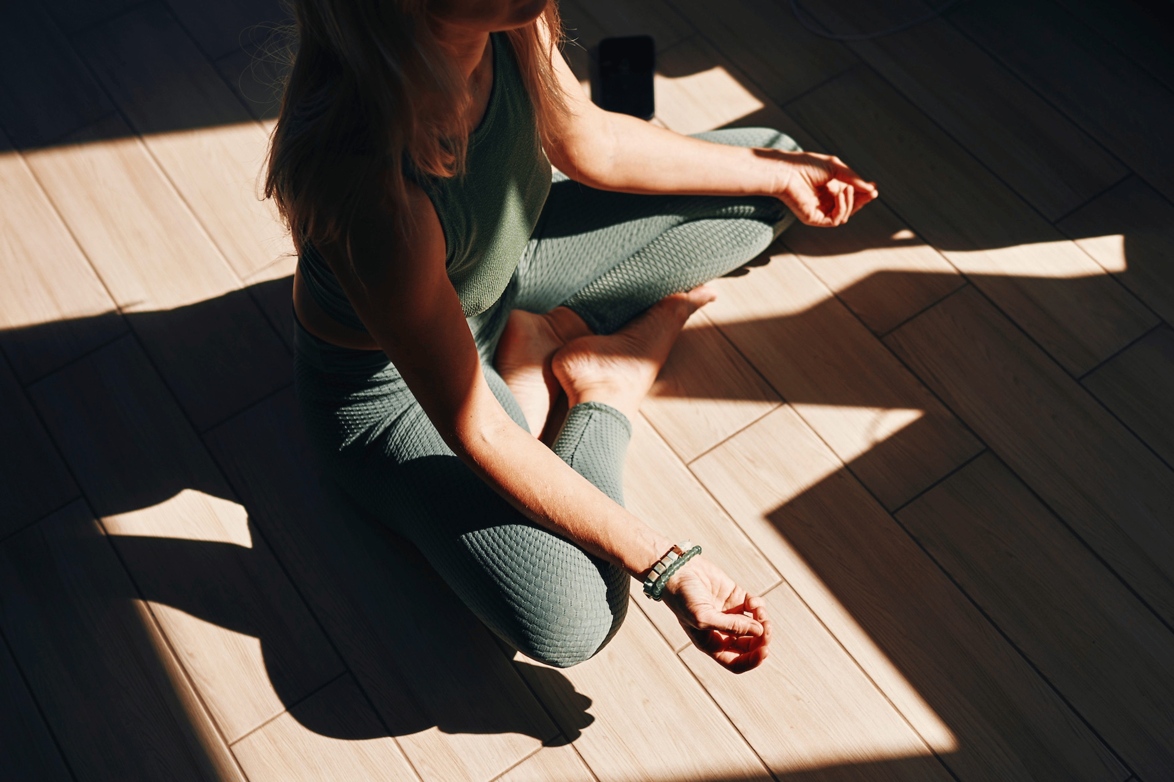 A close-up photo of a woman practicing mantra meditation at home. She's sitting in a criss-cross position with her hands and arms resting on her legs.