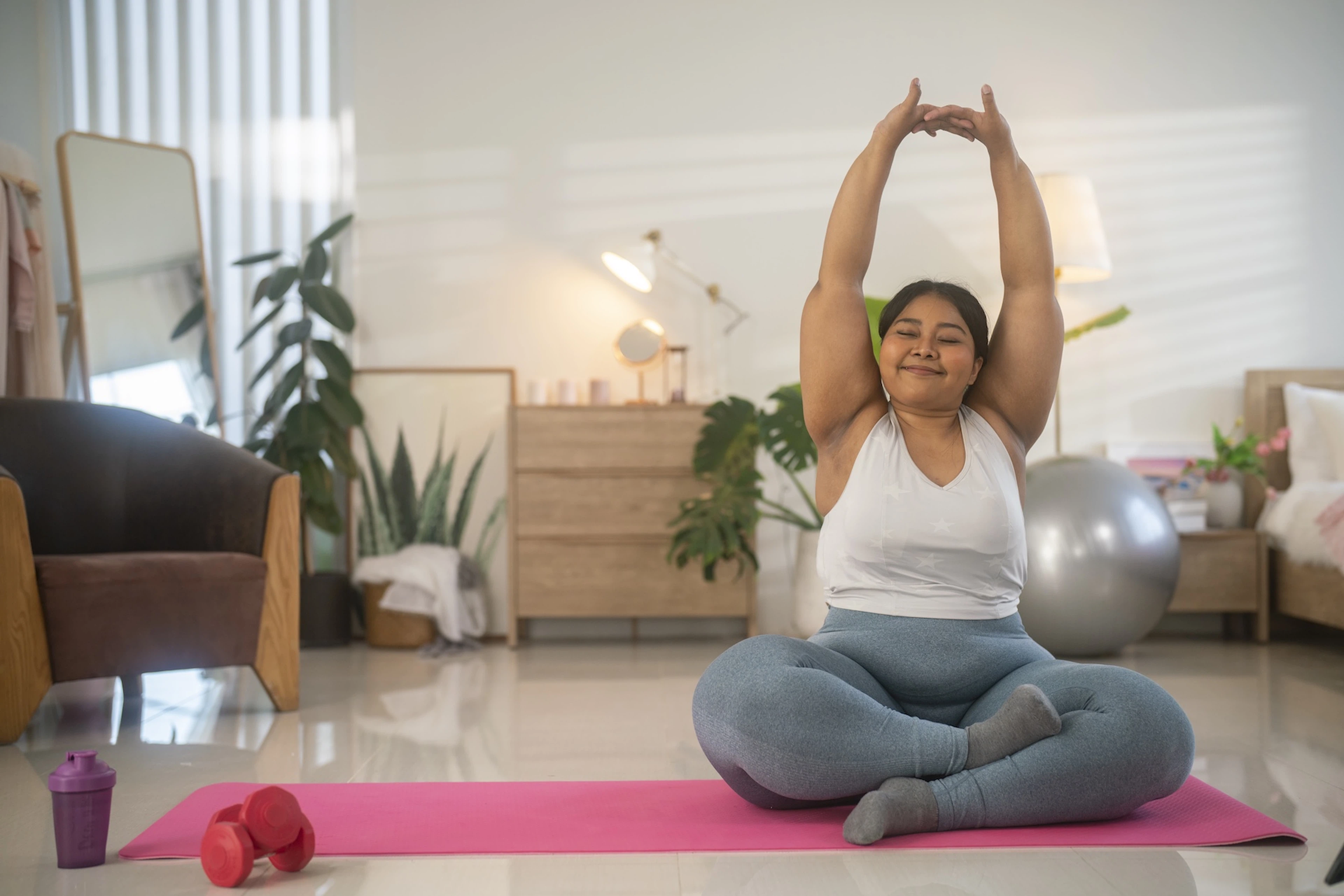 A woman smiling and sitting on her yoga mat at home. Her eyes are closed and her arms are stretched overhead, her hands connected.