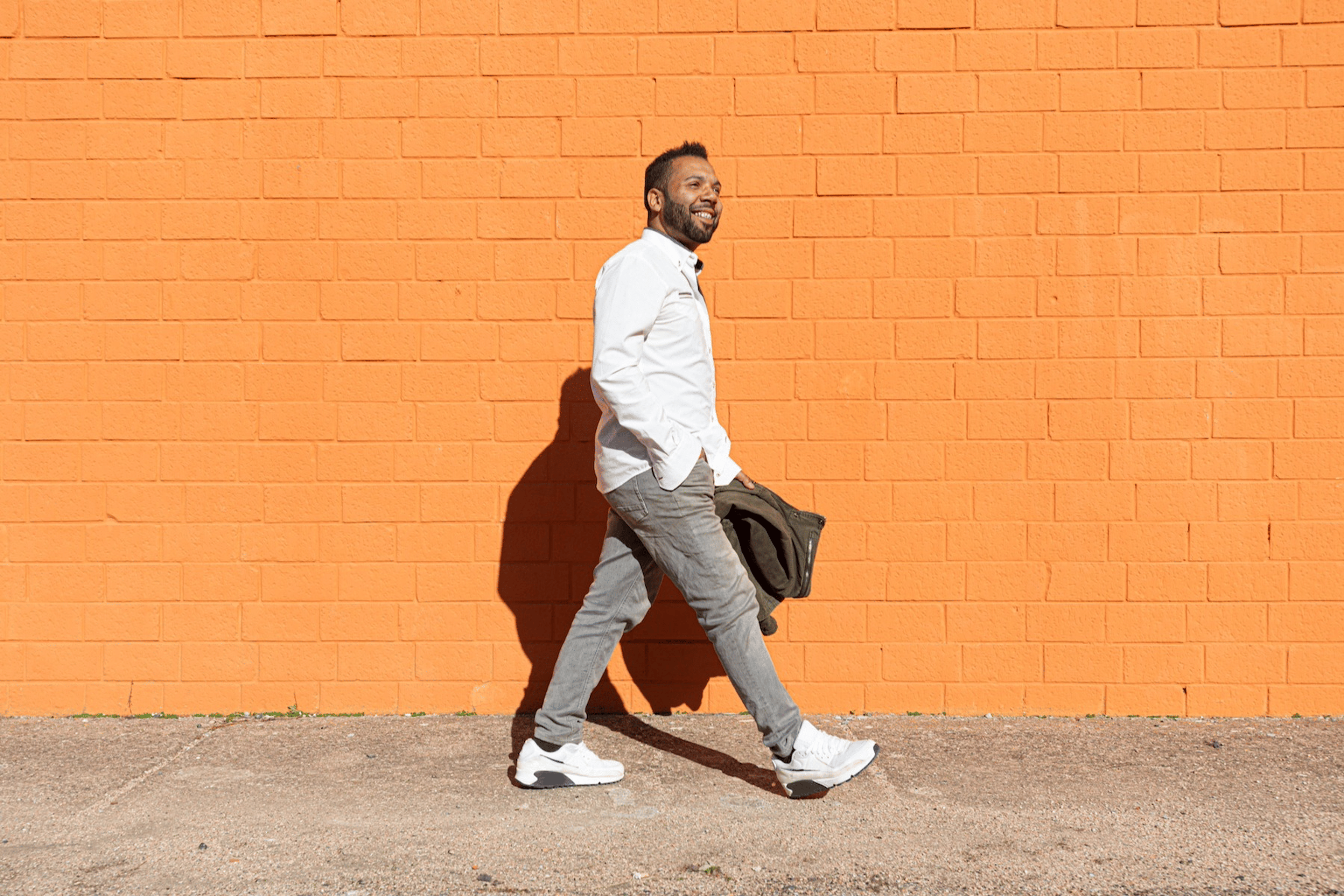 A man going for a walk in the sunshine. He's smiling and walking along on a bright orange brick wall. Learn the mental health benefits of walking in this article.